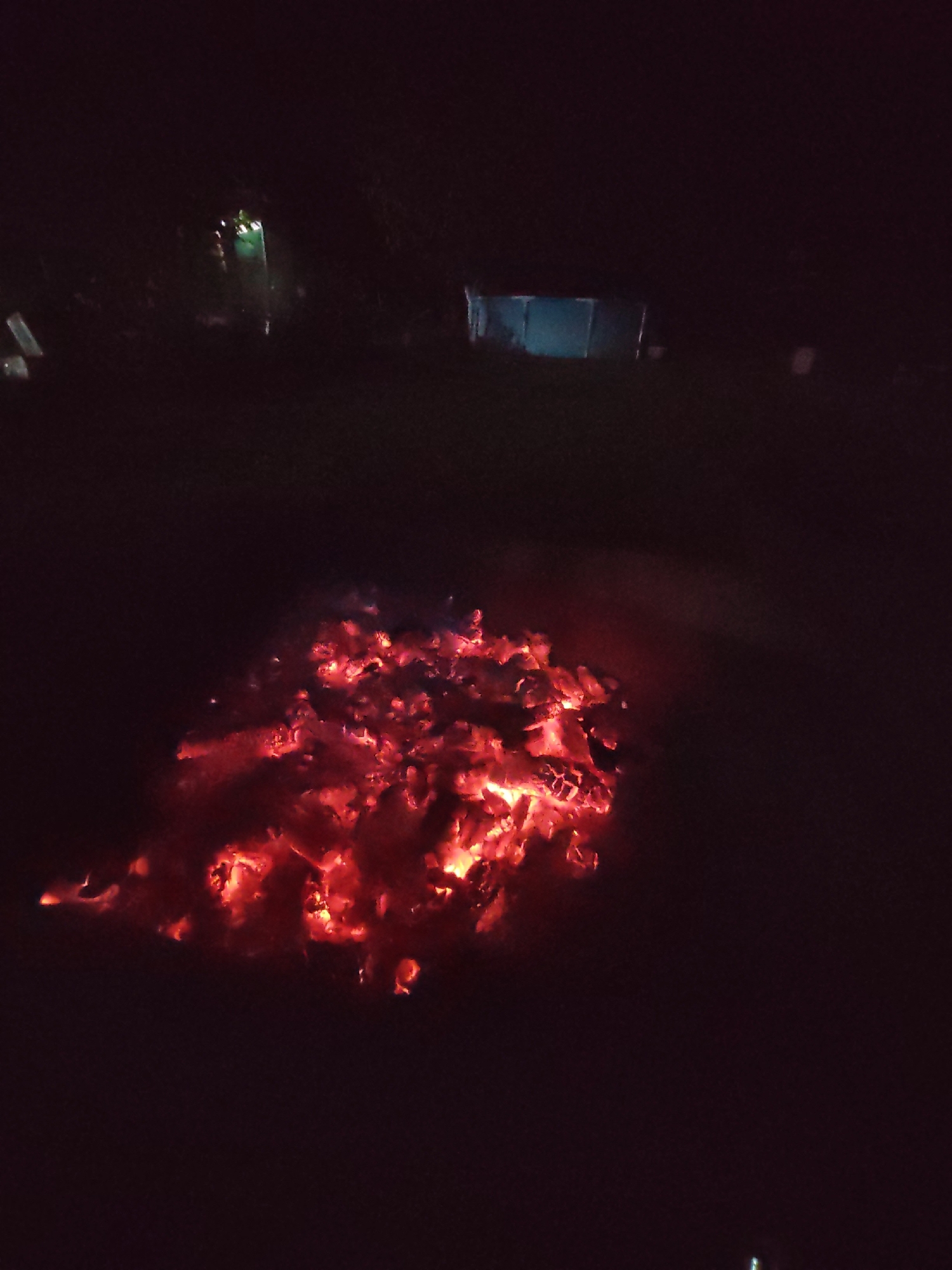 An aerial, high-angle shot captures a pile of glowing red embers in the foreground against a dark, outdoor environment. The embers are concentrated in the lower center of the frame, emitting a bright red and orange light that illuminates the immediate surroundings and casts a faint red haze. In the background, there are indistinct structures that appear to be dimly lit, possibly a building or shed with a blueish light emanating from it on the right, and another object with green light on the left. The overall scene is dark and suggests nighttime conditions. The location in Psebai, Russia, is implied by the context provided, but not visually evident in the image itself.