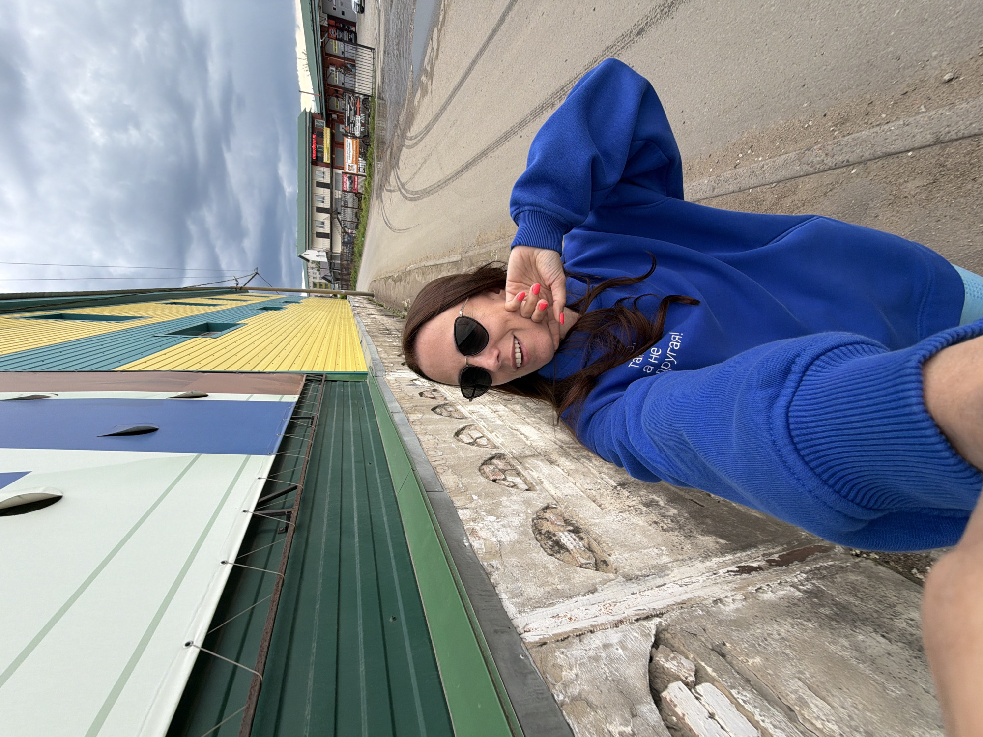 The image is a selfie taken by a woman in an outdoor, urban setting in Birobidzhan, Russia. The woman appears to be lying on her back or leaning against an elevated concrete surface, smiling directly at the camera. She has long brown hair and is wearing a bright blue hoodie and dark sunglasses. Her fingernails are painted a vibrant coral or orange color.
To her left, a building is prominently featured, characterized by its distinct horizontal corrugated metal siding in alternating yellow and blue colors at the top, transitioning to green and white sections lower down. Another part of the same structure or an adjacent one shows solid green siding. The concrete surface she is on is rough and weathered, with some parts appearing broken or worn.
In the background, behind the woman, a concrete path or road is visible, marked with distinctive tire tracks. Further in the distance, several low-rise commercial buildings with various advertisement banners and fences can be seen under a heavily clouded, overcast sky. The overall atmosphere suggests daytime during possibly dull weather.
Notable text includes Russian writing on the woman's blue hoodie, which reads: "Та..., а не другая!" (translating to "This..., and not another!" or "Such..., and not different!"). Some of the distant advertisement banners also feature Russian text, though specific details are largely unreadable from this distance, apart from a potential "автозапчасти" (auto parts). The woman's expression is joyful and relaxed.