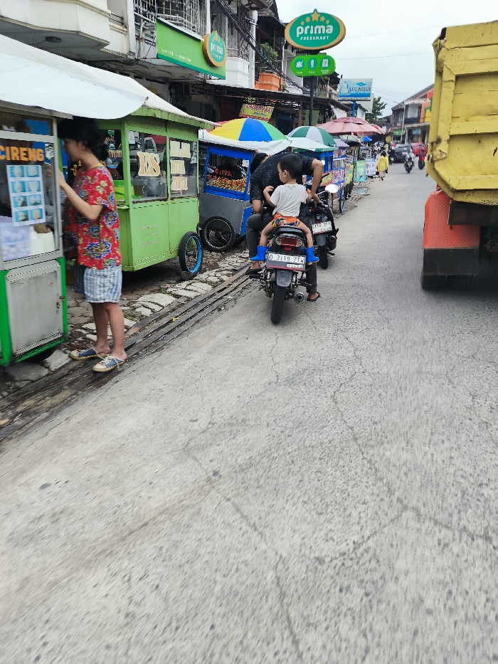 The scene takes place on a street in Kecamatan Dayeuhkolot, Indonesia, at the coordinates -6.963 latitude and 107.594 longitude. Three men and a young boy are visible in the image. The men are all wearing casual clothing. Two of the men are standing in front of food stalls, while the other man is on a motorcycle. The young boy is riding on the back of the motorcycle. The motorcycle is parked in front of a food stall. There are other food stalls and buildings visible in the background. There are some people walking in the background. The street is paved with concrete. The environment is a busy street scene.