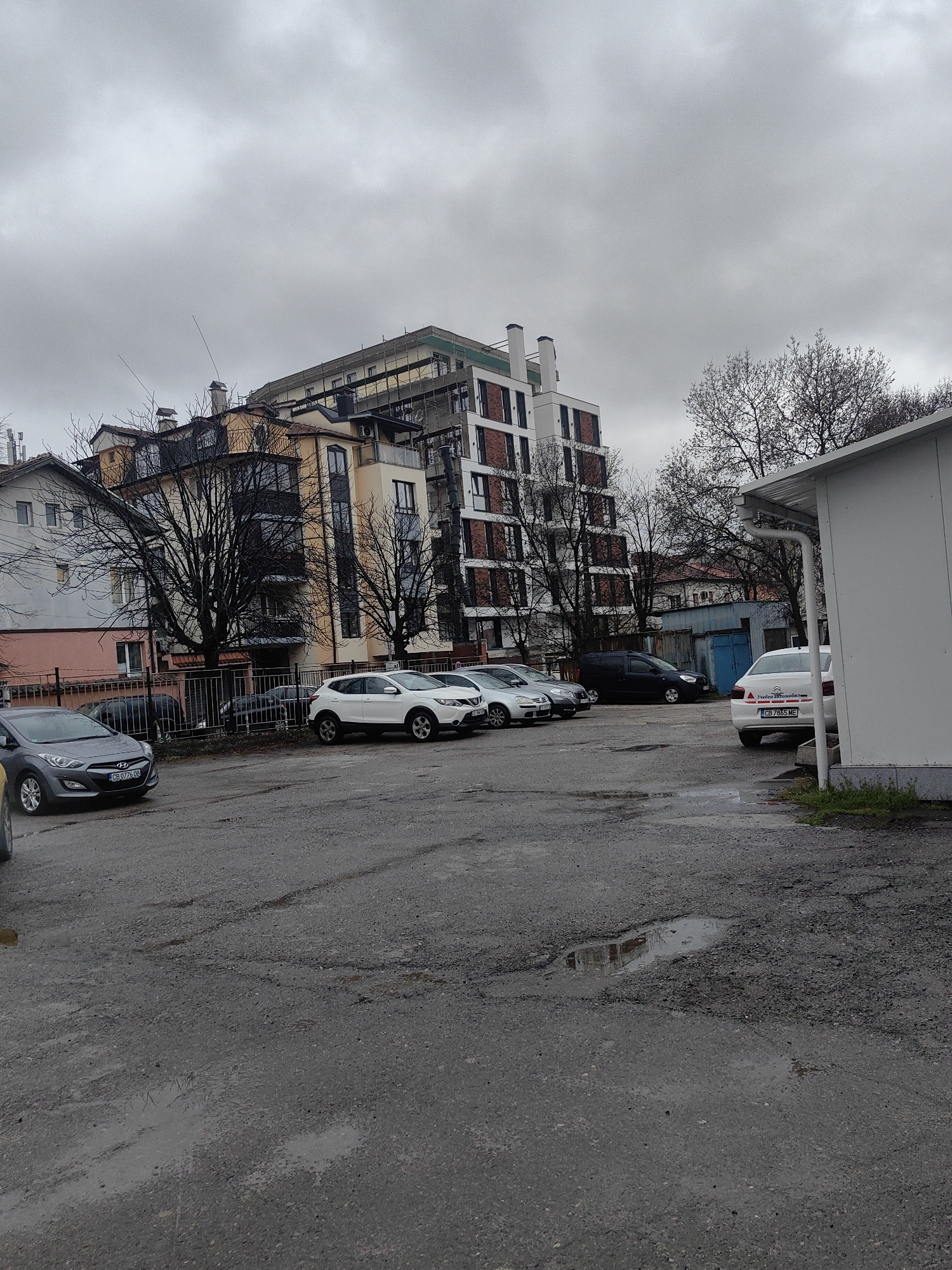 The image depicts an outdoor parking lot in Sofia, Bulgaria on an overcast day. The lot is paved with asphalt that is cracked and contains puddles. Several parked cars are visible, including a gray Hyundai with license plate "CB 0776 OB", a white Nissan Qashqai, and a white Citroen with license plate "CB 7865 ME". In the background, there are several buildings of varying heights and architectural styles. Bare trees are scattered throughout the scene. A white prefabricated structure is partially visible on the right side of the frame. The overall impression is one of urban decay and a lack of maintenance.