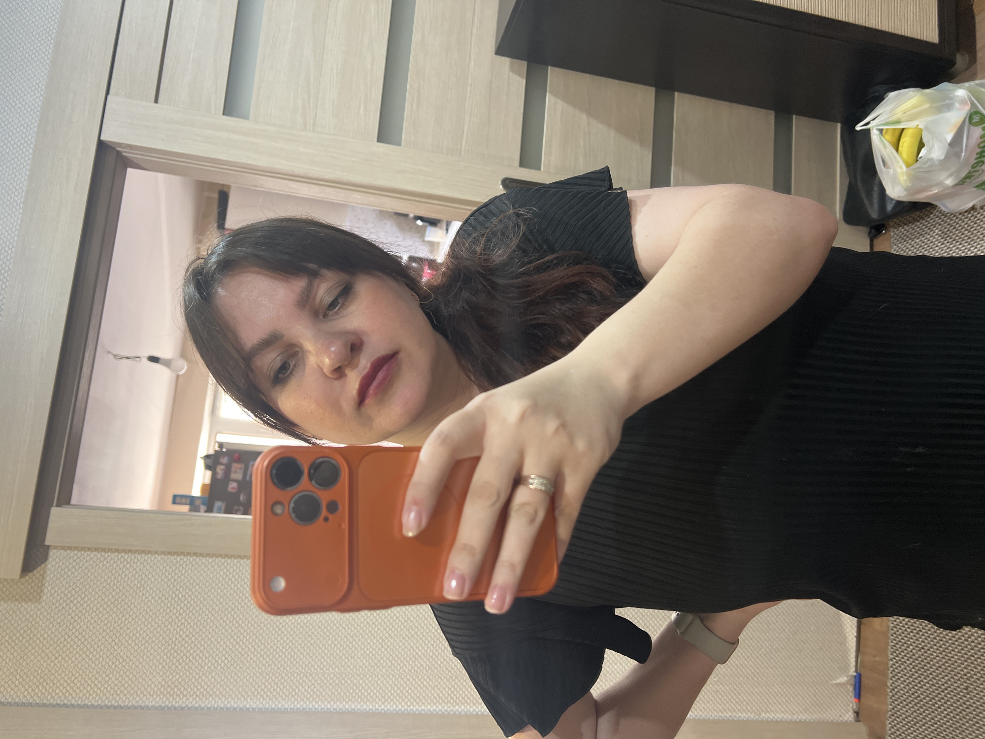 A woman is taking a selfie with an orange phone in front of a mirror. She is wearing a black dress and a ring on her finger. In the background, there is a door, a cabinet, and a bag of bananas. The location appears to be inside a residence. There is no verifiable evidence to indicate that this photograph was taken in the city of Noviy, Russia.
