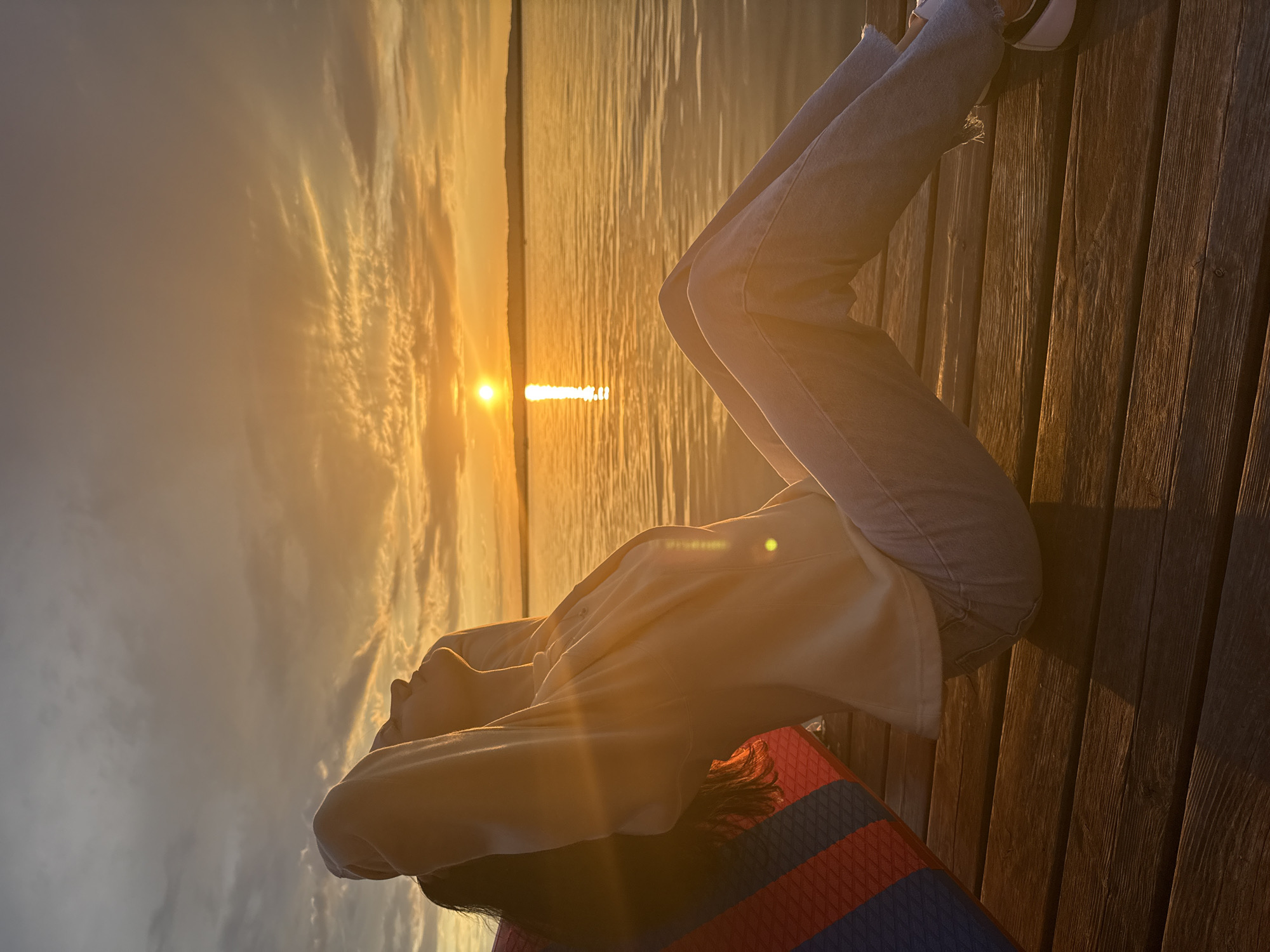 A person is sitting on a wooden pier at sunset, facing the water. The sun is low in the sky, casting a warm golden light across the scene. The water reflects the orange and yellow hues of the sunset, and a gentle ripple is visible on its surface. The person is leaning back, with their arms behind their head, appearing relaxed. They are wearing a light-colored top and jeans. To the left of the person, a colorful object, possibly a cushion or a life vest, is propped against their side. The wooden planks of the pier are visible in the foreground, showing texture and grain. The horizon line is defined by a distant landmass. The sky is filled with scattered clouds, illuminated by the setting sun. This scene is taking place at the Kama River embankment in Chaikovsky, Russia.