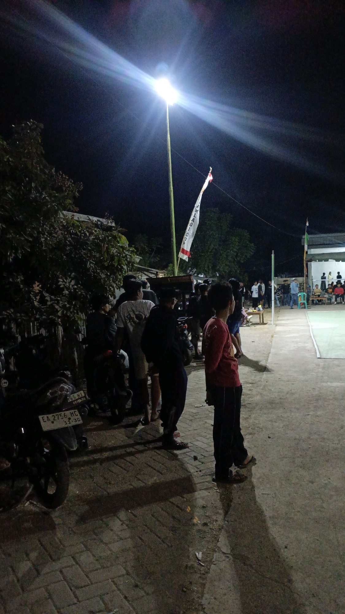 The image depicts an outdoor scene at night in Taliwang, Indonesia, illuminated by a bright overhead light pole. A group of people, mostly young males, are gathered in the foreground and midground. They appear to be standing around and observing an event or activity taking place in the background, which seems to be a volleyball court with spectators seated on benches.
In the immediate foreground, a motorcycle is parked on a brick-paved area. Shadows are cast on the ground from the light source and the people. The environment suggests a casual gathering, possibly for a sports event. The flag visible in the midground is white with red lettering. The ground surface transitions from brick paving to a more dirt-like or sandy texture near the court. The overall atmosphere is informal and community-oriented.