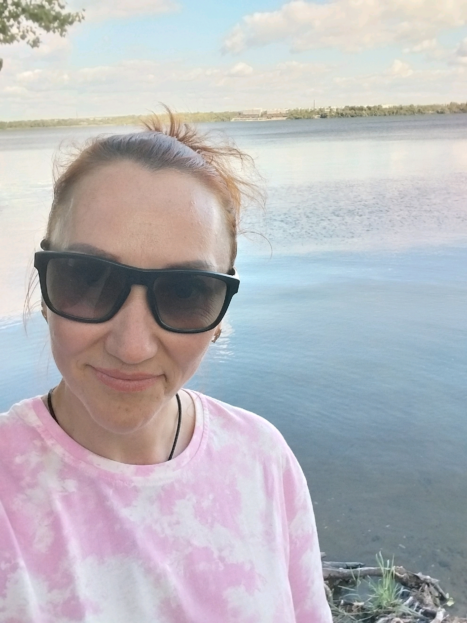 A woman is taking a selfie by the Dnipro River in Dnipro, Ukraine. She has her hair pulled up in a messy bun and is wearing large black sunglasses and a pink and white tie-dye t-shirt. She is smiling slightly and looking at the camera. Behind her, the river stretches out to the horizon, with a city skyline visible in the distance. The sky is partly cloudy. There is a small pile of driftwood and vegetation on the right side of the frame.
