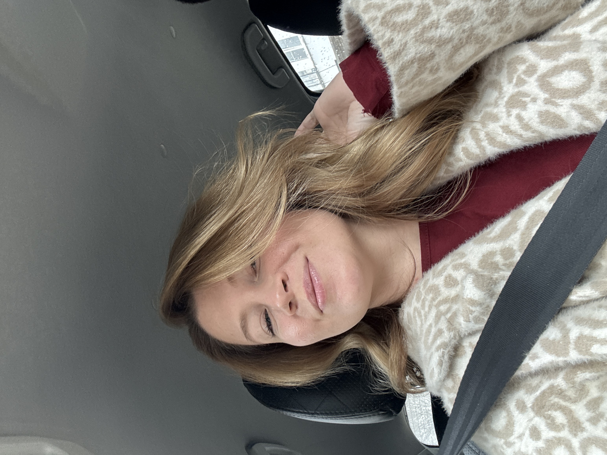 The image captures a woman inside a car in Chaikovsky, Russia, likely seated as a passenger. She has long, wavy light brown hair and looks directly at the camera with a subtle, closed-mouth smile. She is wearing a dark red top and a fuzzy, cream-colored cardigan with a light, abstract patterned design, possibly leopard print. A black seatbelt crosses diagonally over her chest. Her right hand is visible near her head, gently touching her hair.
The setting is the interior of a car, with a black headrest and a dark gray ceiling visible. Through the window on the left side of the frame, a blurred exterior view suggests urban architecture and wet weather, with visible droplets on the glass, indicating rain or snow. The natural lighting is diffused, suggesting an overcast day. The woman appears relaxed and content. No visible text is present in the image.