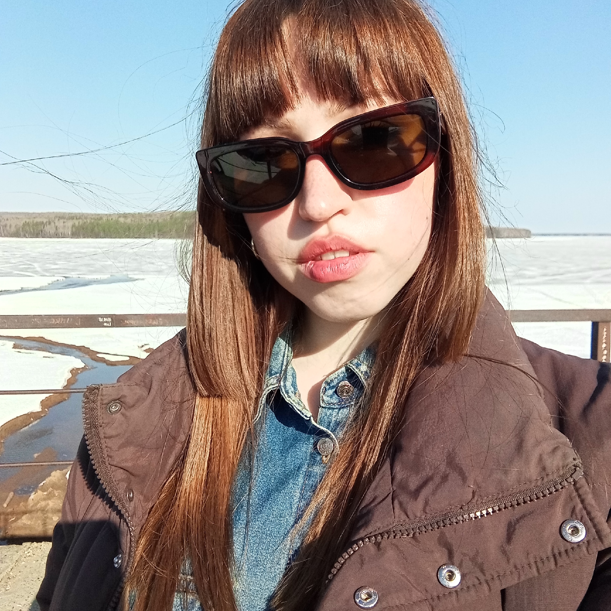 A young woman wearing sunglasses, a denim shirt, and a brown jacket takes a selfie outdoors. The background appears to be a frozen body of water. The location is most likely the city of Chaikovsky, Russia. The presence of ice suggests it may be spring or winter.