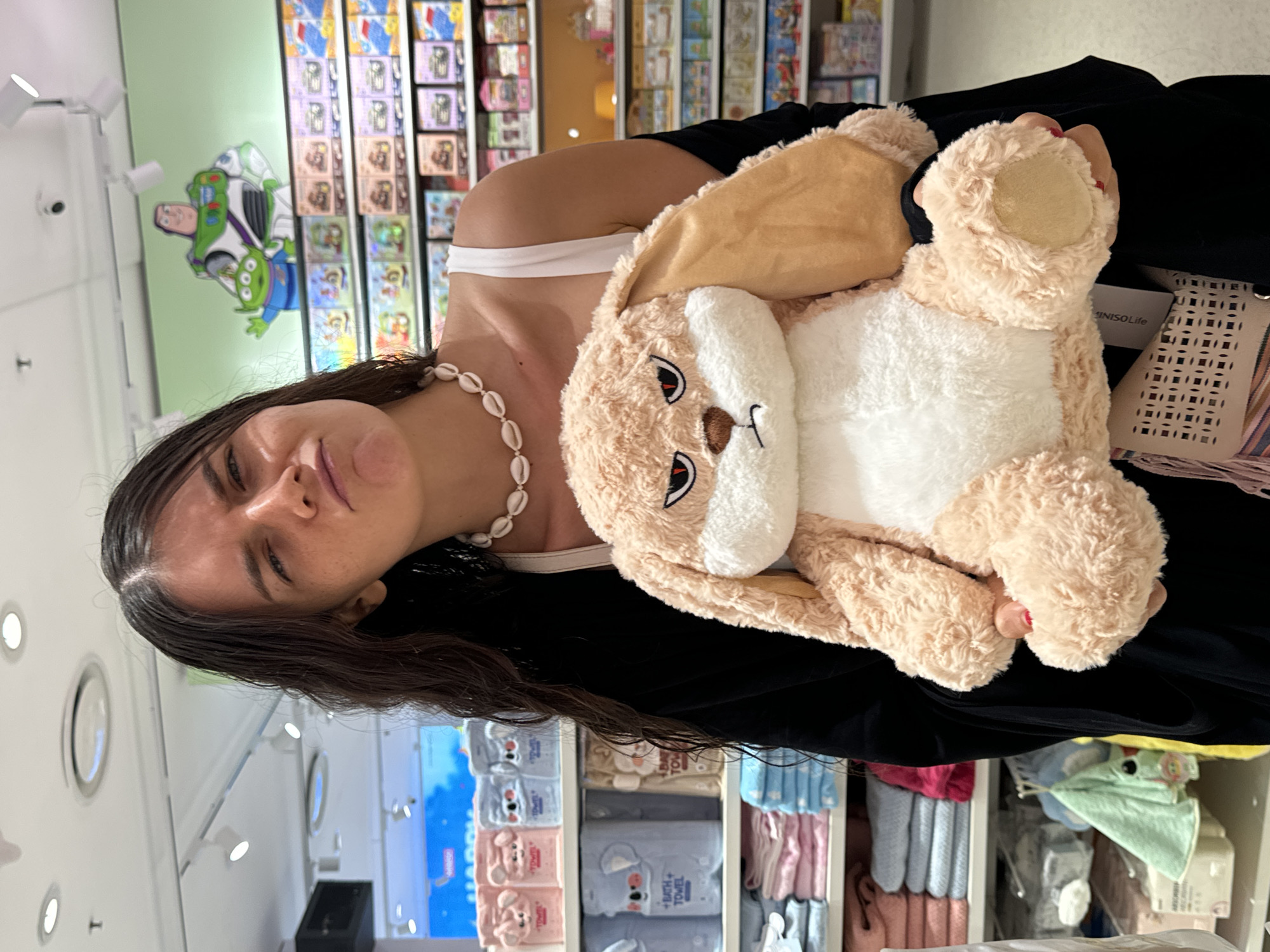 A young woman with dark, wavy hair is holding a large, light brown, fuzzy stuffed rabbit with an angry expression. She is wearing a white tank top and a black cardigan, and a seashell necklace. Her facial expression is a pout or grimace. Behind her, shelves are stocked with various items, including folded towels in shades of pink and blue, and on the top shelf, there are boxes of merchandise. A large mural of Buzz Lightyear and an alien from the Toy Story franchise is visible on the wall above the shelves. The lighting appears to be from overhead fluorescent lights. The overall setting suggests a retail environment, possibly a department store or a toy store. The location is not specified as Phu Thuy, Vietnam.