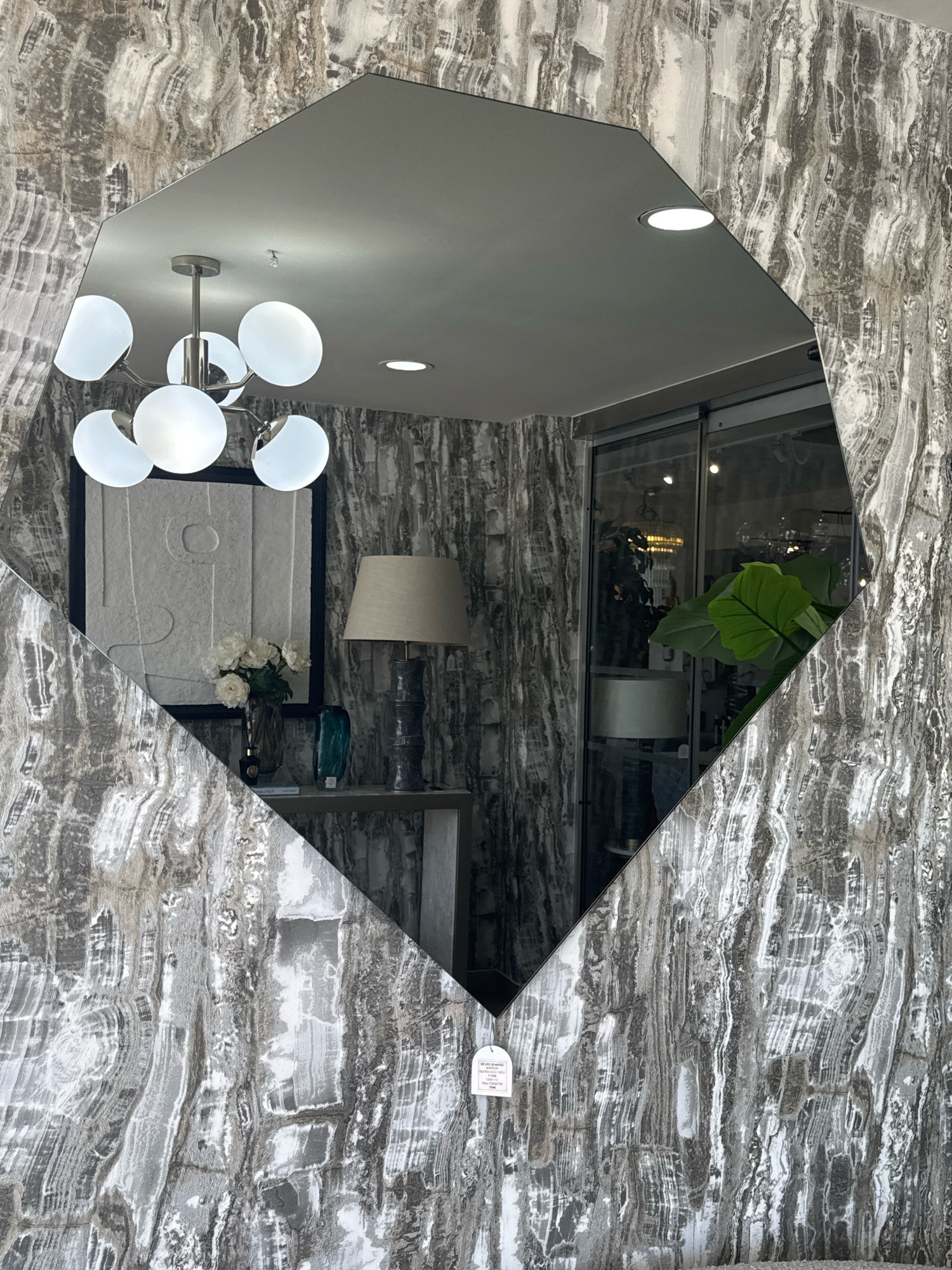 A large, irregularly shaped mirror with a beveled edge is centered in the frame, reflecting a portion of an interior. The mirror is mounted on a wall covered in a striking gray and white marble-patterned wallpaper. The reflected scene shows a contemporary chandelier with multiple globe-shaped lights, a recessed ceiling light, and a glimpse of a doorway or large window with glass doors. Below the chandelier, a framed piece of abstract art hangs on the reflected wall, accompanied by a vase of white flowers, a dark blue vase, and a table lamp with a beige shade on a console table. To the right of the reflected scene, a large, leafy plant is visible through the glass doors, hinting at the space beyond. A white tag with black text is hanging from the bottom edge of the mirror. The mirror is angled, revealing the marble wallpaper on either side of it.