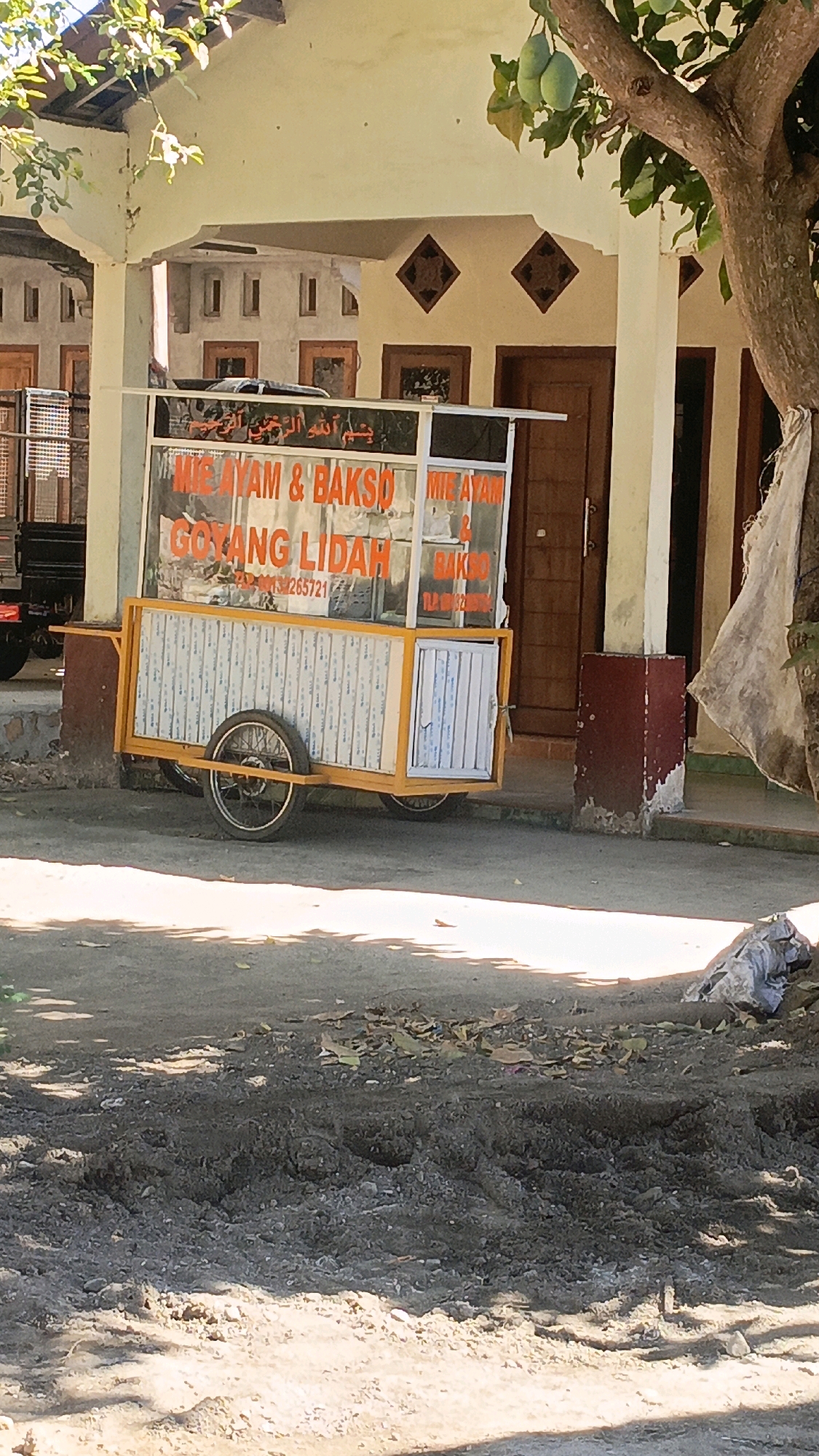 The image depicts a food cart parked on an unpaved area in Taliwang, Indonesia. The cart, which appears to be yellow and white, displays signage advertising "MIE AYAM & BAKSO GOYANG LIDAH" and "MIE AYAM & BAKSO". A portion of a building with arched openings is visible in the background, and a tree with green fruit hangs over the scene. The ground in the foreground is rough and uneven, with scattered leaves and debris, suggesting a somewhat undeveloped or construction-affected area. Sunlight casts shadows across the scene. There are no people visible in the image.