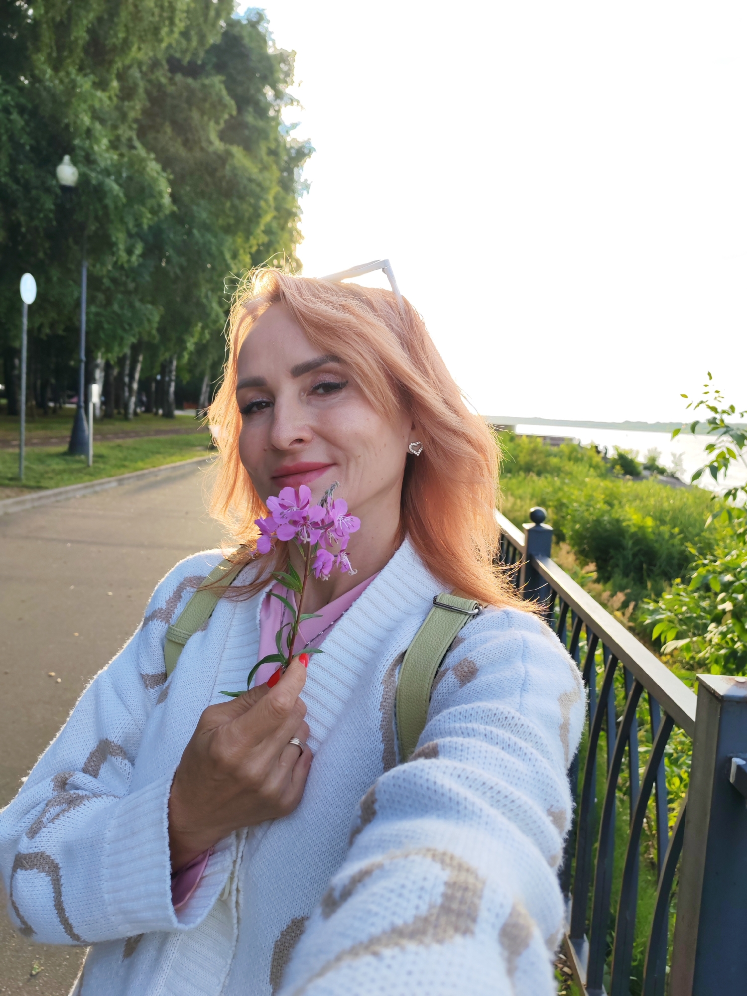 A middle-aged woman with orange-tinted hair is holding a small purple flower and taking a selfie outdoors. She is wearing a white knitted cardigan with a beige patterned design over a pink top. A light green backpack strap is visible on her shoulder. She has on earrings, makeup, and a ring on her finger. The background shows a park with trees and a path, and a railing along a body of water, suggesting a location like the embankment in Bol'shie Mkhi, Russia. The lighting suggests it is late afternoon or early evening.