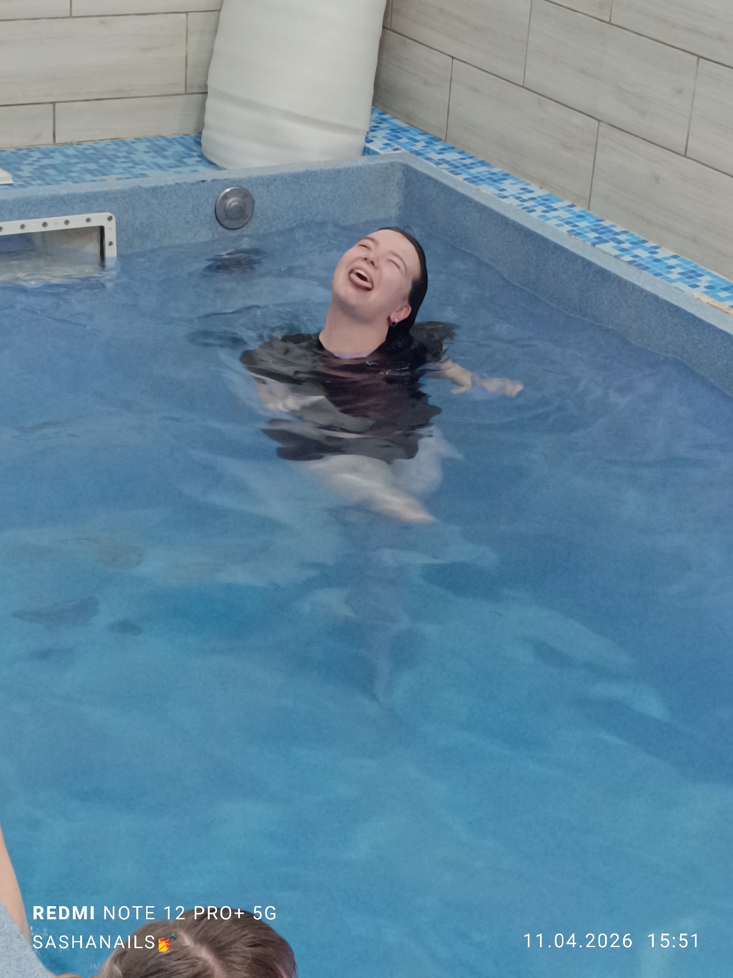 A person is in a pool filled with water. The pool is blue, and there's a white, cylindrical object leaning against the edge of the pool. Tiles are visible along the edge and wall of the pool area. The person is partially submerged, with their head above water. They are looking upwards with their mouth open. The date and time "11.04.2026 15:51" are visible in the bottom right corner of the image. A portion of another person's head is visible in the bottom left corner. The location is Chaikovsky, Russia.