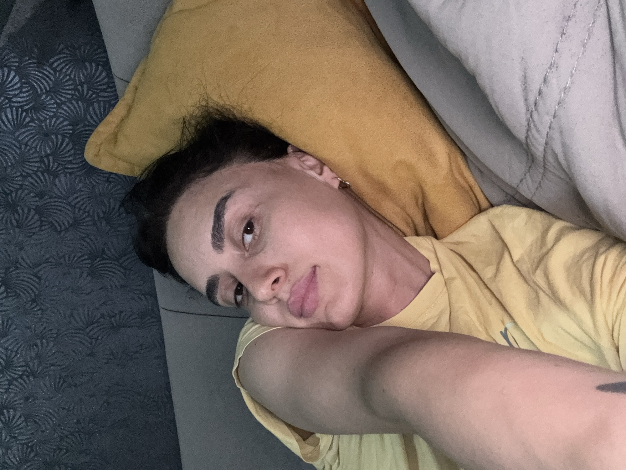 A person is lying on a couch with a yellow pillow behind their head. The person is wearing a yellow t-shirt and has their arm resting on their face. The person is looking at the camera. The couch is in a room with patterned wallpaper. The image was taken in Tbilisi, Georgia.