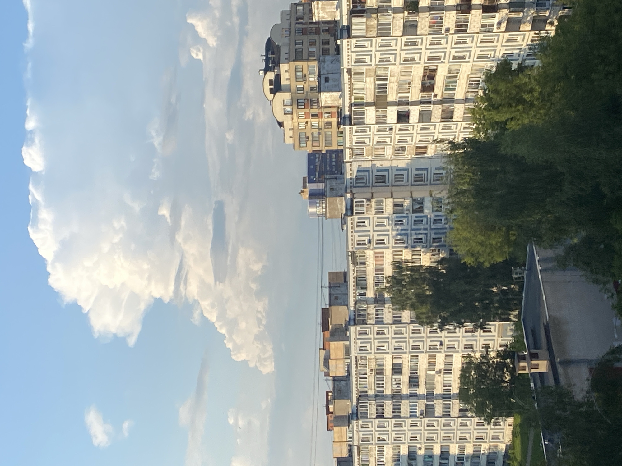 The image depicts a view of multi-story residential buildings in the city of Dubna, Russia, under a partly cloudy sky. The buildings are typical of Soviet-era architecture, characterized by their modular design and numerous windows. Several trees are visible in the foreground and between the buildings, indicating a green, urban environment. In the background, a large, fluffy cloud formation dominates the sky, illuminated by sunlight. The scene appears to be captured during the daytime, possibly late afternoon or early evening due to the warm light. There are no people or specific actions visible in the frame, focusing solely on the architectural and natural elements of the cityscape.