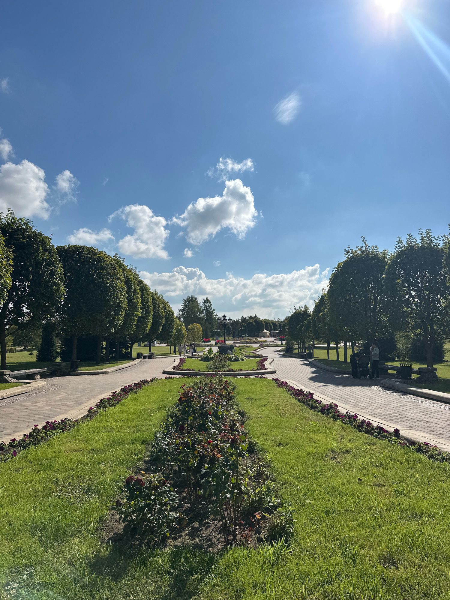 The image depicts a park scene in Minsk, Belarus, on a sunny day with scattered clouds. In the foreground, a manicured lawn with flowerbeds lines a paved pathway. Beyond the pathway, a row of evenly pruned trees creates a symmetrical border. Further in the distance, more trees and park features are visible, including lampposts and a fountain-like structure in the center of a circular garden area. Several people are seen walking and standing on the pathways, some in small groups. The sky is bright blue with the sun high in the upper right corner, casting light and shadows across the scene.