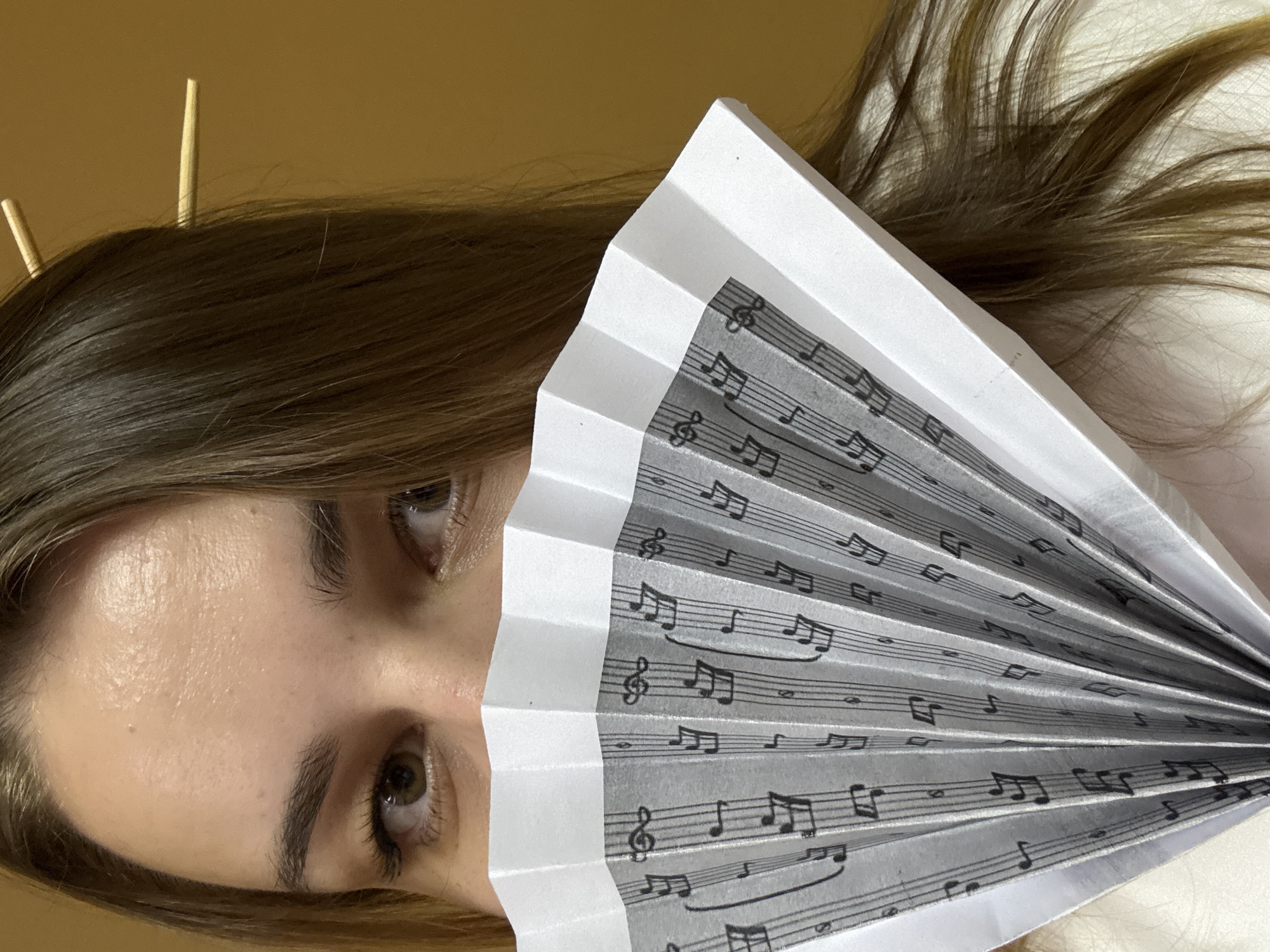 The image displays a close-up, vertically oriented portrait focusing on a person's upper face and a decorative fan. The subject, likely female, has light brown hair that flows around the visible parts of their face. Two golden, slender hair sticks are inserted into the hair on the left side of the frame.
The person's eyes, appearing brown or hazel, are open and gazing in a neutral direction. The forehead and area around the eyes are clearly visible, with a clear complexion. The lower half of the face is obscured by a partially fanned-out, white paper fan. This fan is adorned with black musical notation, including treble clefs, staff lines, and various notes and rests, printed across its pleated surface.
The background behind the person's head is a plain, light brown or tan wall. To the right, behind the fan, there is a soft, light-colored surface, possibly a pillow or clothing. The lighting is soft and even, suggesting an indoor setting.
The image captures a posed moment, perhaps an artistic or conceptual shot, without indicating a specific activity beyond the subject holding the fan to their face. There are no strong emotional expressions evident. The only visible "text" consists of the numerous musical symbols and staff lines on the fan. Although the location context is given as Chaikovsky, Russia, there are no specific visual cues within the image itself to confirm this geographical detail.