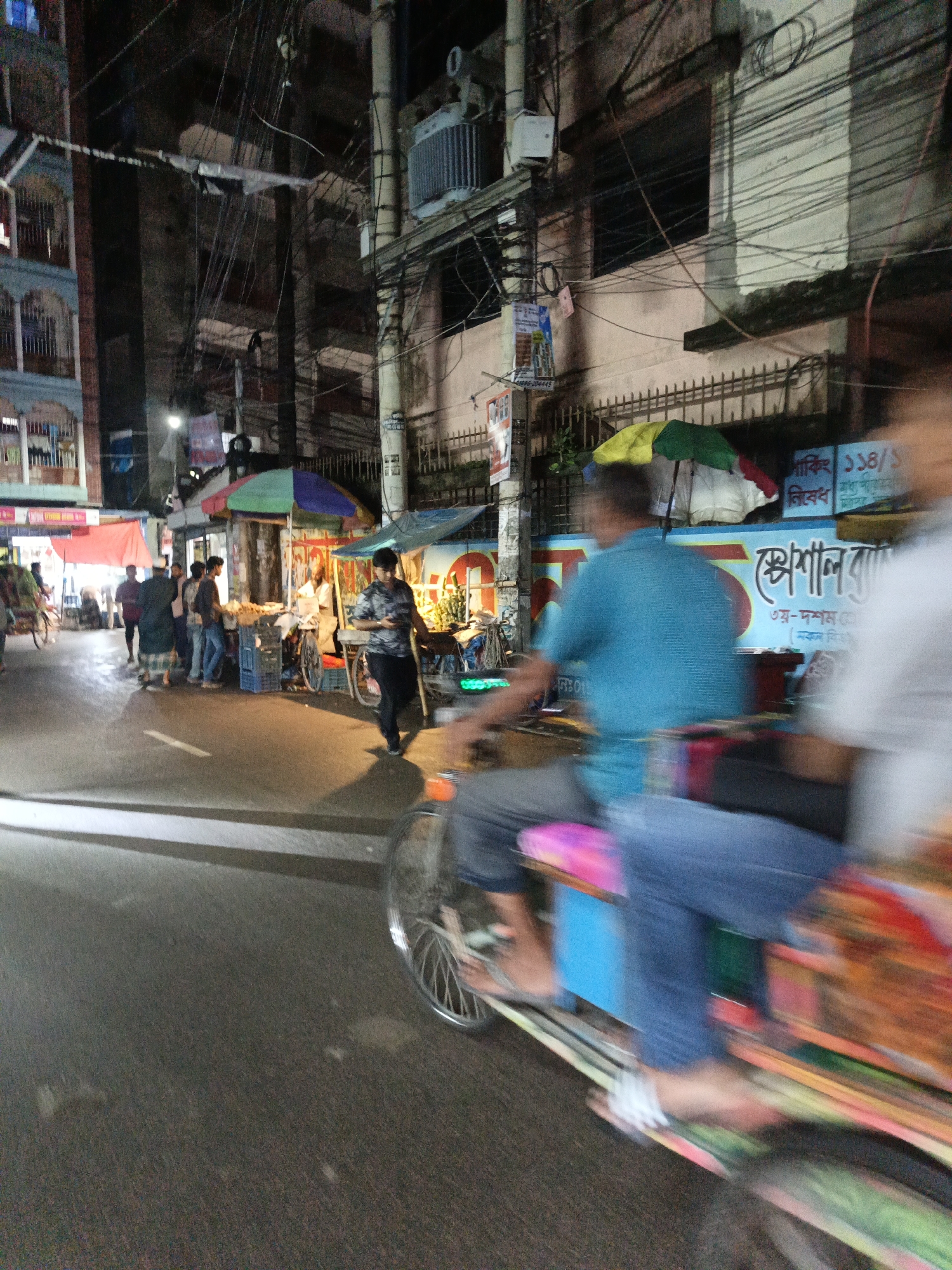 The image depicts a street scene in Dhaka, Bangladesh, at night. In the foreground, a rickshaw is in motion, blurring the figures of its occupants and the vehicle's colorful decorations. The rickshaw is on a paved road with white lane markings. Behind the rickshaw, a man in a blue shirt and jeans is walking or standing. Further back, a bustling market scene unfolds. Several people are gathered around small stalls, some under umbrellas. One stall has a man in a checkered lungi and a blue shirt. Another stall is selling goods under a colorful umbrella, with items like fruits and vegetables visible. Buildings line the street, adorned with numerous electrical wires. Signs in Bengali are visible on the buildings and stalls. The overall atmosphere suggests a lively and active urban environment at night, typical of Dhaka.