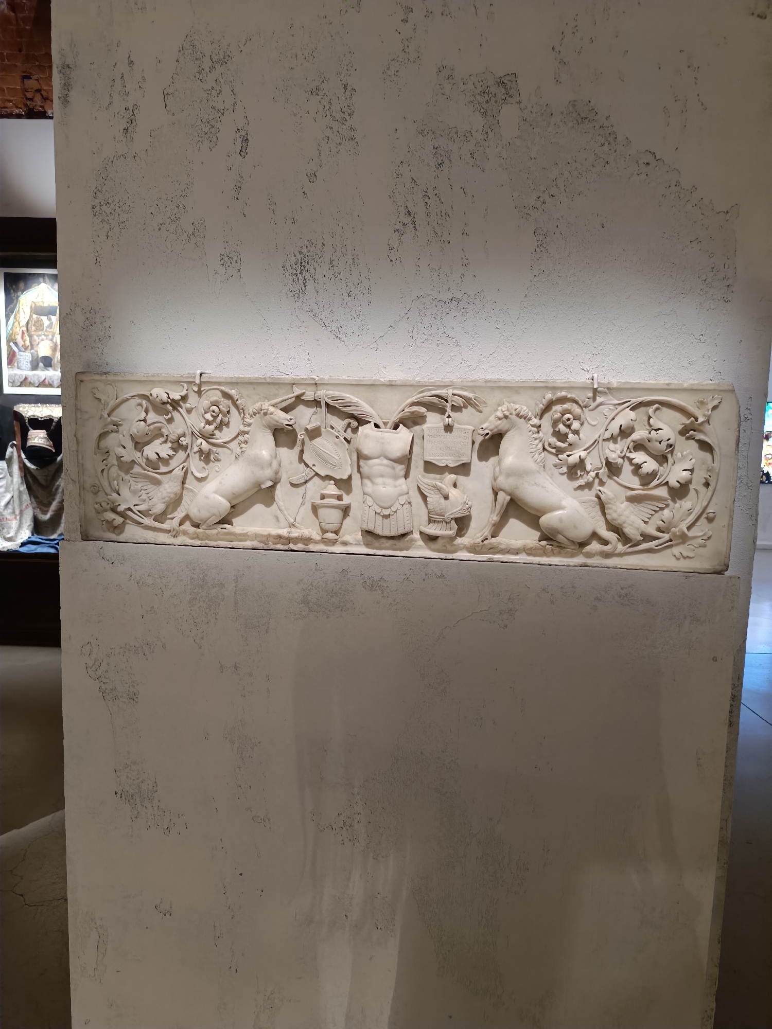In an indoor setting with textured white walls, a long, white marble relief is mounted horizontally. The relief depicts an intricate scene with mythological or allegorical figures. On the left, a horse is depicted in mid-rear, with stylized foliage and a bird to its left. In the center, a torso of a muscular figure, possibly a warrior or god, is shown, adorned with armor and a shield. To the right of the torso, another horse is depicted in a similar dynamic pose, facing left, with more foliage and a bird. The relief is framed by decorative elements and has inscriptions visible on a plaque near the center-left. The background on the left side of the image shows a glimpse of other exhibits, including framed artwork and draped fabric. The lighting casts shadows, highlighting the depth and detail of the carving.