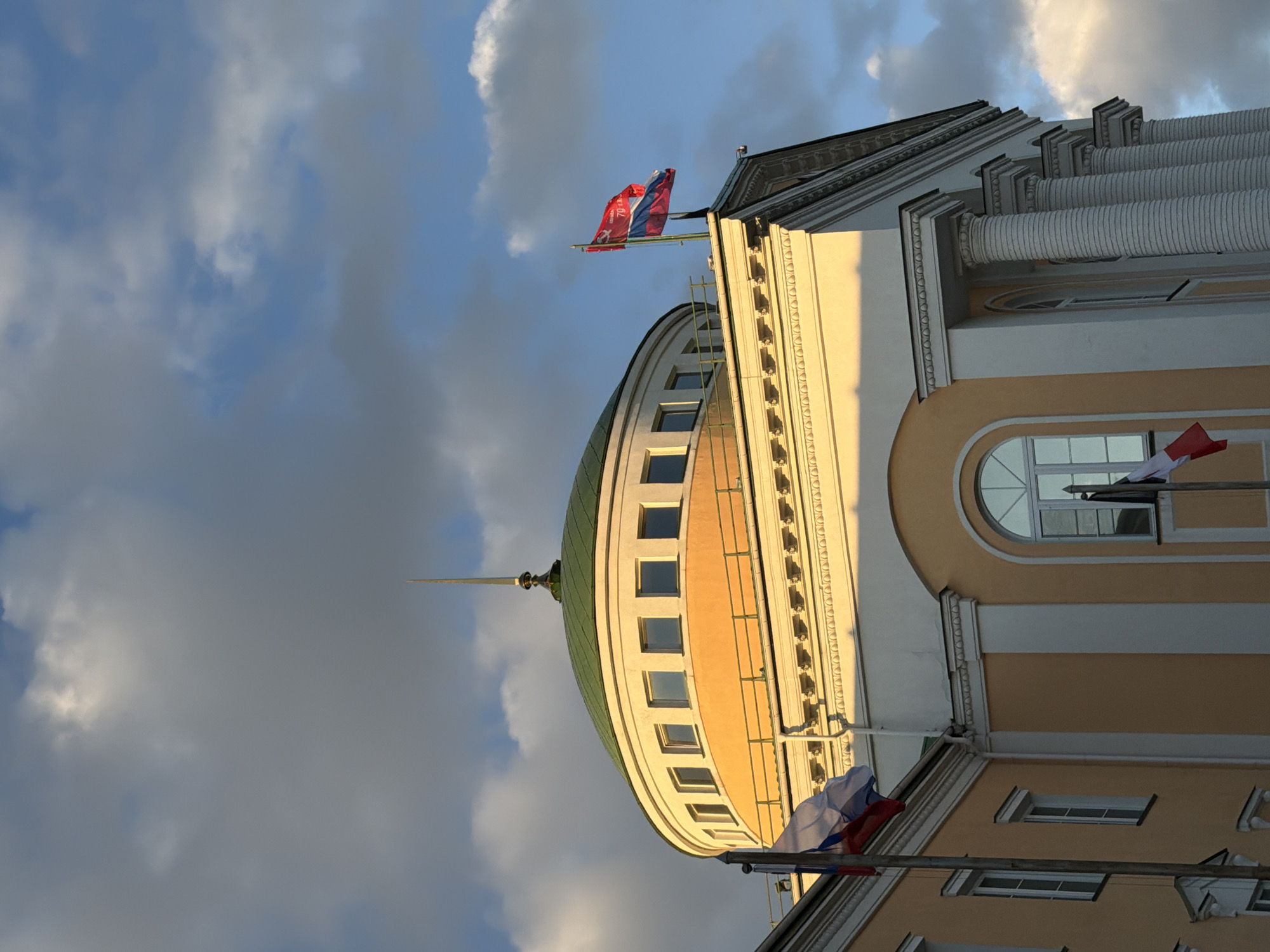 A low-angle, outdoor shot captures the architectural details of a building against a cloudy sky. The building features a rounded dome with a green roof, framed by rows of windows and decorative elements. Adjacent to the dome, the building's facade includes white columns, arched windows, and sections painted in a warm, yellow hue. Several flagpoles protrude from the building, displaying flags with red, white, and blue colors, hinting at a possible national or governmental association. The sky behind the building is filled with a mix of light and dark clouds, suggesting either early morning or late afternoon lighting conditions. While the image does not explicitly pinpoint the location, its architectural style and the presence of the flags suggest a landmark in Izhevsk, Russia.