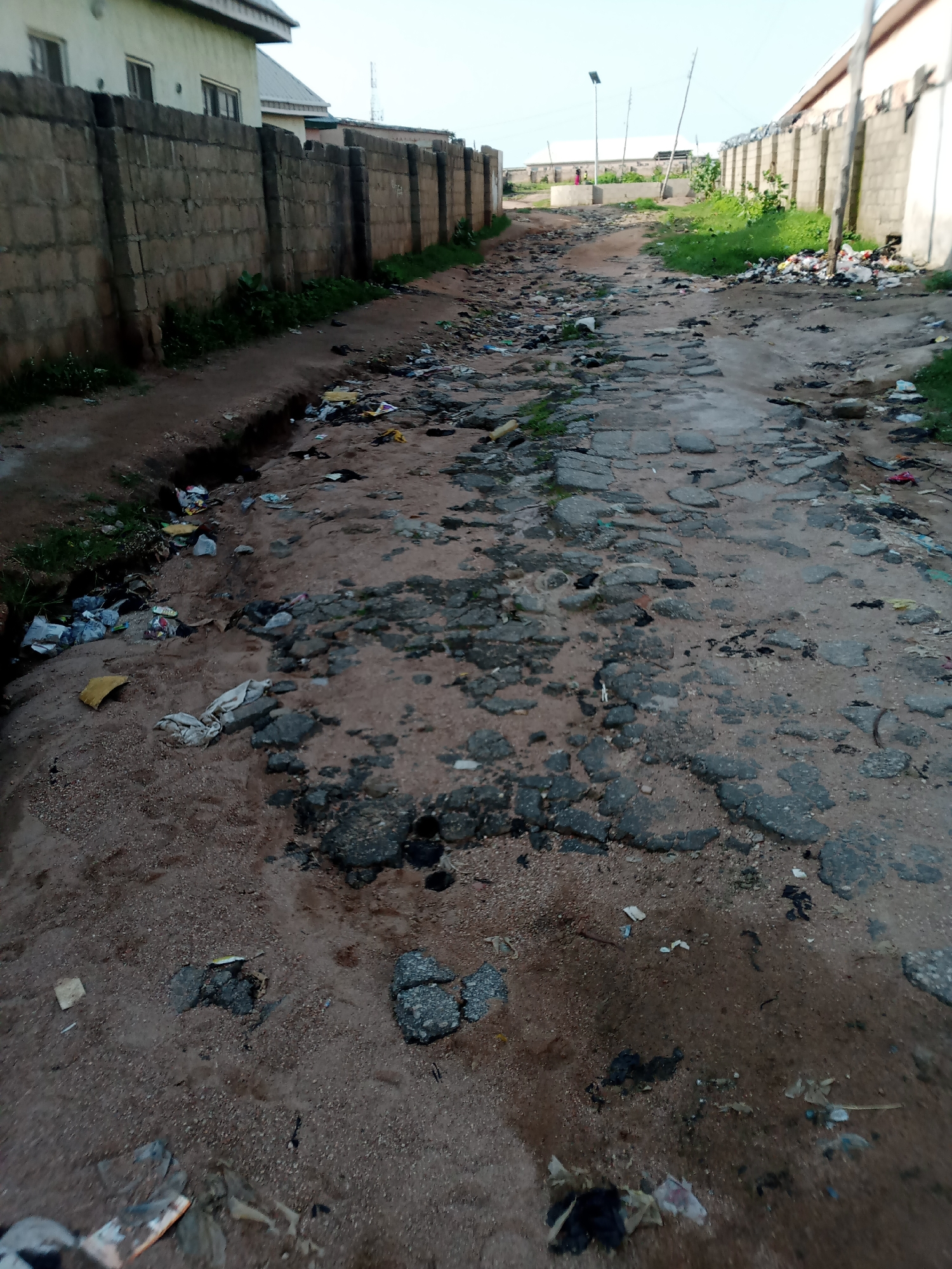 The image shows a severely deteriorated road in Jauro Yinu, Nigeria. The road surface is broken and uneven, with large patches of missing asphalt and exposed dirt and gravel. Debris, including plastic bags, wrappers, and other refuse, is scattered widely across the road and its shoulders, indicating a significant littering problem. The road is bordered on the left by a tall, stone block wall, behind which buildings with light-colored walls and dark roofs are visible. On the right side of the road, a similar wall is present, with some vegetation growing alongside it and a large pile of trash accumulating near the wall. In the distance, the road continues, appearing similarly unkempt and littered. The sky is overcast, suggesting a dim or possibly rainy day. No people are clearly visible or engaged in any activity within the frame. The overall impression is one of neglect and poor infrastructure.