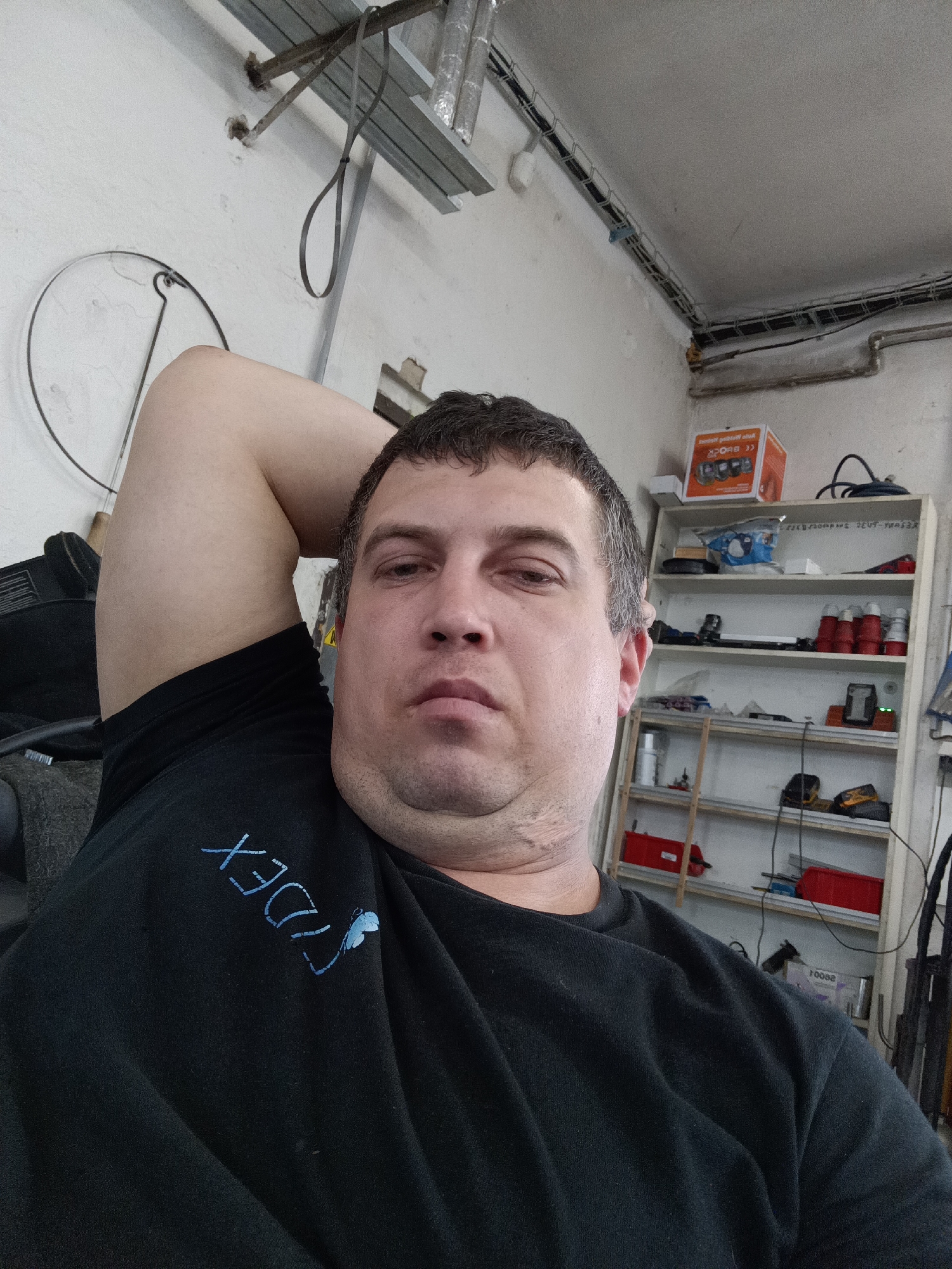 A man is captured in a close-up, chest-up selfie, looking directly at the camera with a neutral expression. He appears to be in his 30s or 40s, with short dark hair and dark eyes. His left arm is raised and bent, with his hand resting behind his head, giving a relaxed pose. He is wearing a black t-shirt or sweatshirt. On the left chest area of his shirt, there is light blue embroidery. The visible text reads "XEDλY" or "XEDTY" (with 'λ' being a stylized letter or symbol), along with a small blue, butterfly-like symbol next to it.
The setting appears to be a workshop, garage, or utility room in Dalovy, Czechia. The background features light-colored walls with visible wear and tear. Exposed metal framing for a ceiling or utilities is visible in the upper left, from which a gray strap hangs. Wires and conduits run along the top perimeter of the walls. On the left mid-ground, a large circular metal frame, possibly a part of an old appliance or antenna, is mounted on the wall.
To the right of the man, a multi-tiered wooden or metal shelving unit is filled with various items. The top shelf holds an orange box with partially visible text (including "auto..." and a "CE" logo) and a coiled black cable. The lower shelves are stocked with miscellaneous tools, red industrial-style electrical plugs, white bottles, and other containers. A yellow and black power tool is also visible on one of the shelves. The room is lit by artificial light, and the overall impression is that of a functional, utilitarian space.