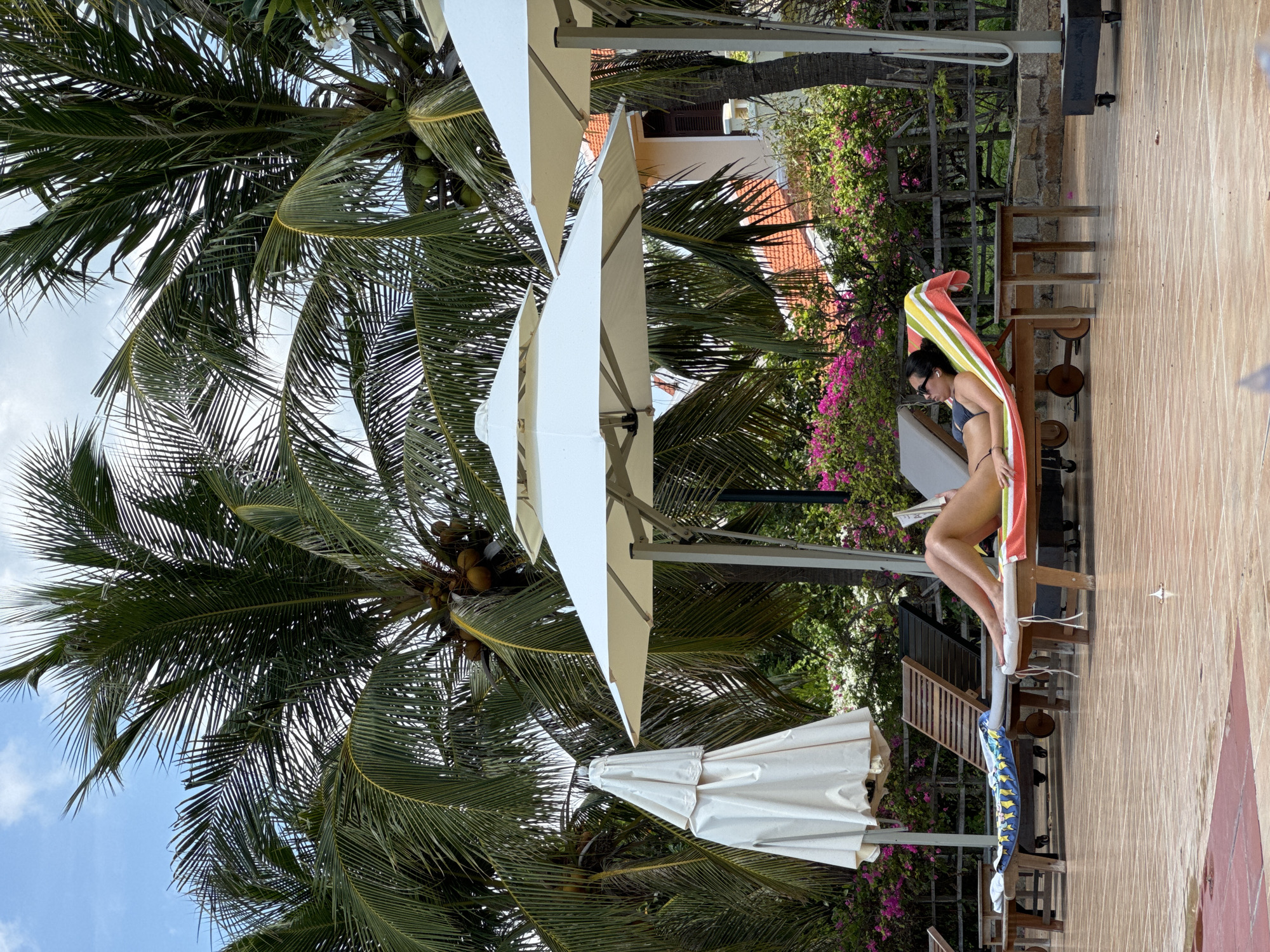 The image shows a woman in a dark bikini seated on a wooden lounge chair next to a swimming pool in Phu Thuy Ward, Vietnam. She is partially covered by a striped towel draped over her shoulders. A smaller patterned towel lies on another chair next to her. The area is shaded by several white parasols and palm trees, with a building visible in the background behind a bougainvillea-covered fence. The pool deck is tiled, with sunlight reflecting off its surface.