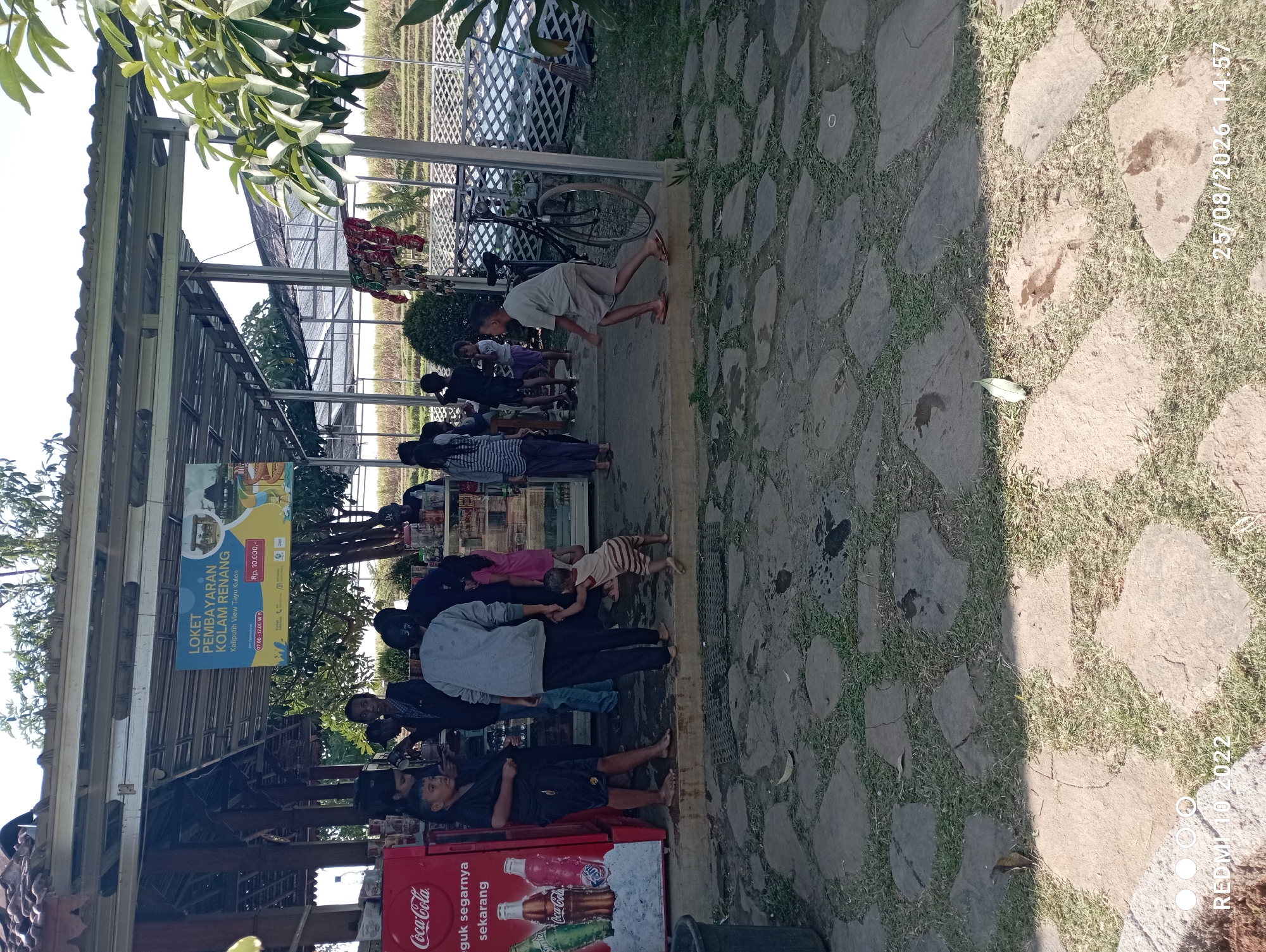 The image shows an outdoor scene at a swimming pool facility in Tayu, Indonesia. A Coca-Cola branded refrigerator is visible on the left. Several people, including adults and children, are gathered around a counter where tickets are being sold for "KOLAM RENANG" (swimming pool). The ticket sign indicates prices and operating hours. In the background, a rice field or cultivated land is visible. There are also bicycles and other objects suggestive of an outdoor recreational area. The ground is paved with irregular stones. The overall atmosphere appears to be casual and sunny, with a sense of everyday activity.