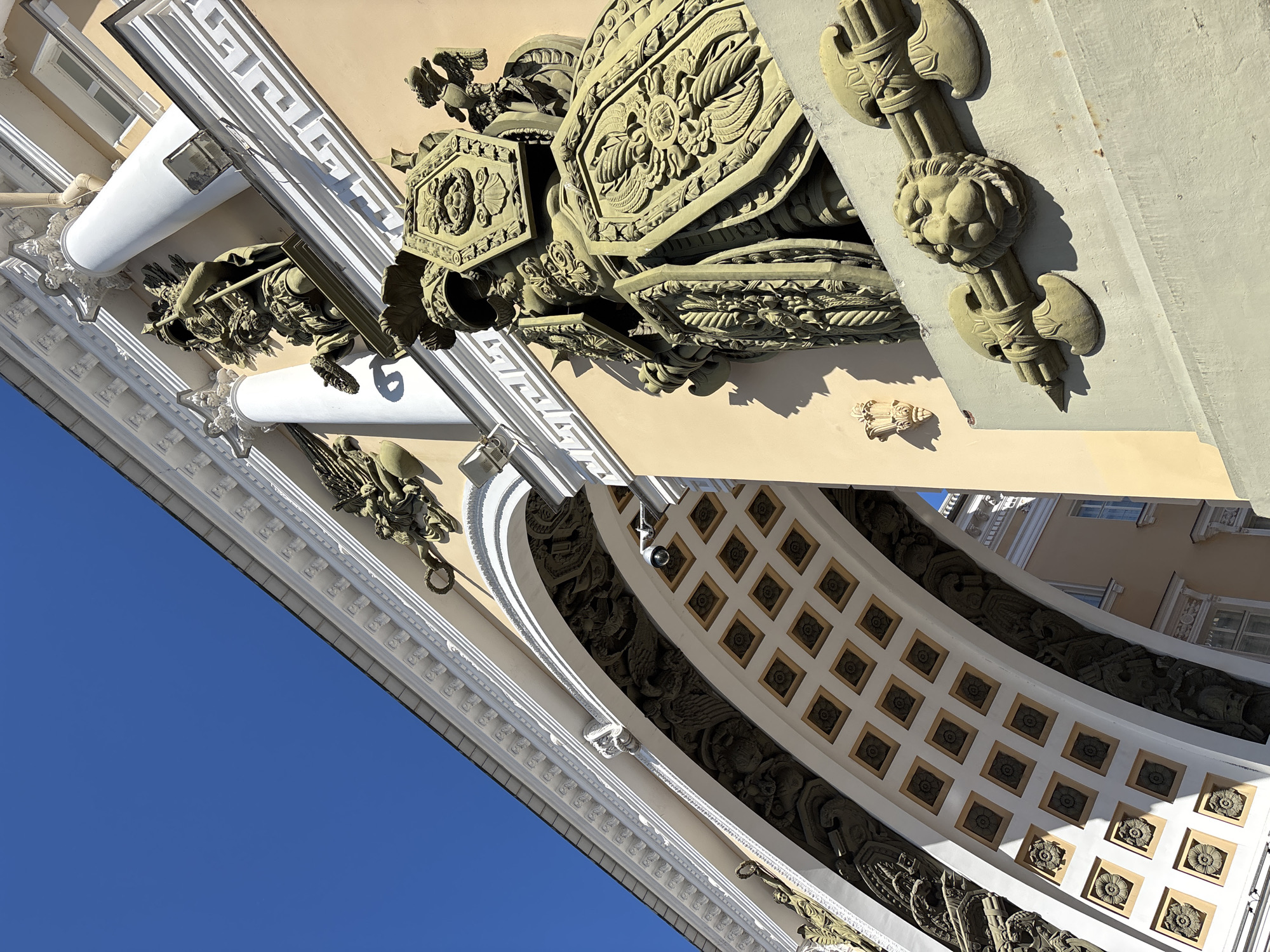 The image captures a low-angle, close-up view of the ornate architectural details of the General Staff Building's archway in Saint Petersburg, Russia. The archway is adorned with numerous sculptures, including a large, heavily armored figure holding a shield and a spear, positioned to the right of the arch. This figure is surrounded by other intricate carvings of mythical creatures and military regalia, such as axes and lion heads. The arch's interior is decorated with a coffered ceiling featuring square panels with floral motifs. A security camera is visible mounted on the arch's surface. The building's facade is painted a pale yellow, contrasting with the white architectural elements and the deep blue sky. The bright sunlight casts strong shadows, highlighting the three-dimensional nature of the sculptures.