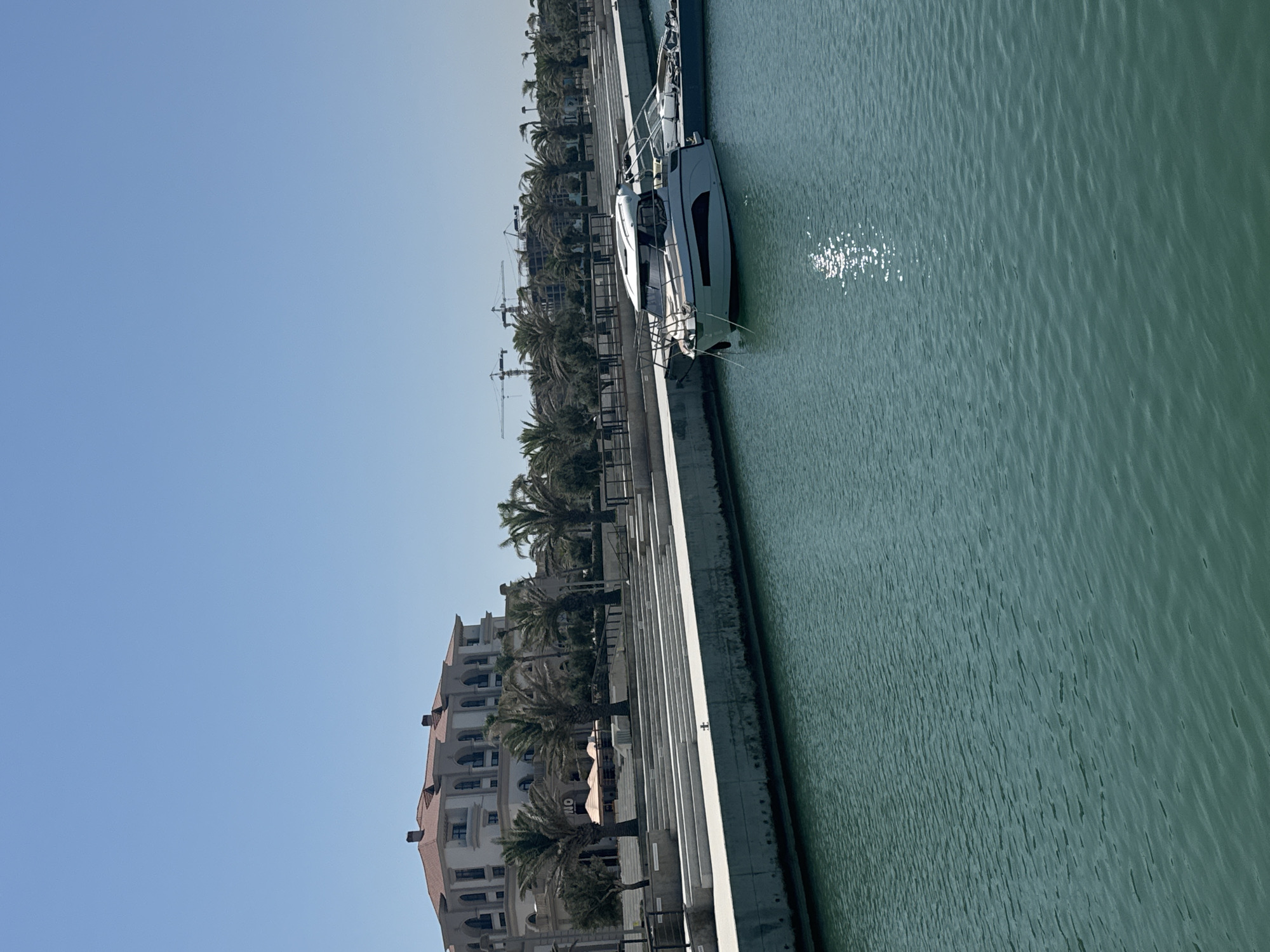 The image depicts a marina area in Sabunchu Raion, Azerbaijan. In the foreground, the tranquil, turquoise water of the marina fills the lower portion of the frame. A modern, white motor yacht with dark accents is moored to the right, its hull reflecting the sunlight on the water. Beyond the yacht, another partially visible vessel is also docked.
Along the waterfront, a tiered promenade with wide, white steps leads up to a street lined with palm trees. Behind the palm trees, a large, multi-story building with a red-tiled roof and white facade stands prominently. The architecture suggests a hotel or residential complex. Construction cranes are visible in the distance, indicating ongoing development in the area. The sky above is a clear, bright blue, characteristic of a sunny day. The scene is devoid of people.