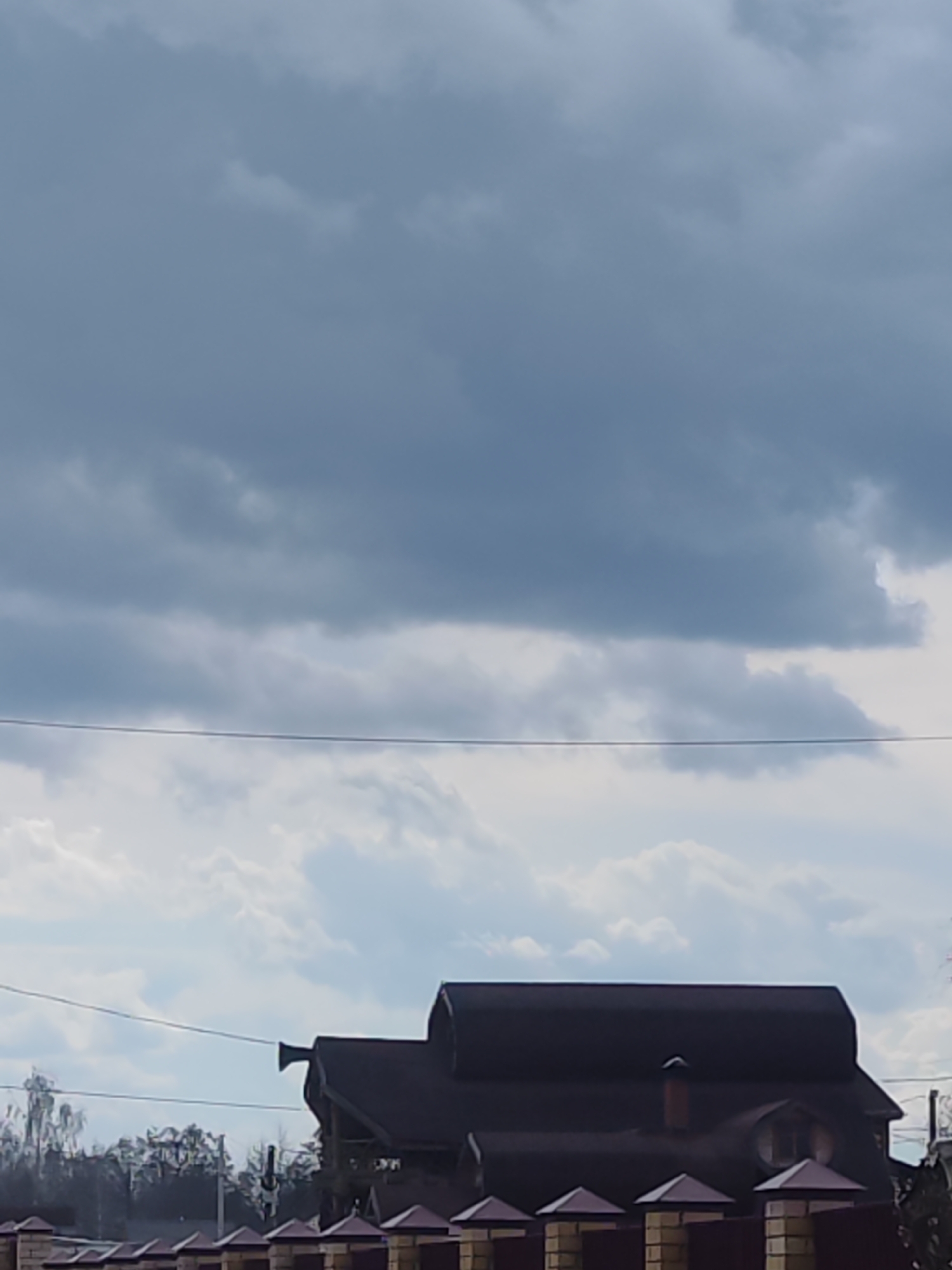 The image captures a static outdoor scene, focusing on a cloudy sky above a house and fence. The upper two-thirds of the frame are dominated by a dense, overcast sky, featuring dark grey clouds in the upper portion and lighter, more scattered clouds below. A single, dark power line stretches horizontally across the middle of the sky, subtly dividing these cloud layers.
In the lower third of the image, the dark roof of a substantial house is visible. The house has a multi-faceted roof design, a small chimney on the right side, and a rounded dormer window. On the left side of the roof, a distinctive, horn-shaped element, likely a ventilation or exhaust pipe, protrudes upwards. Below the house, a fence is prominent, constructed with light-colored, possibly stone or brick, pillars that are topped with dark, pyramid-shaped caps. Dark panels fill the spaces between these pillars. To the left and behind the fence, a cluster of bare trees with thin branches is visible against the lighter clouds.
The scene depicts a cloudy, possibly overcast day, suggesting muted light rather than direct sunlight. No people or specific activities are taking place; it's a still landscape or architectural shot. There is no visible text. The setting appears to be suburban or rural, consistent with the location context of Chaikovsky, Russia.