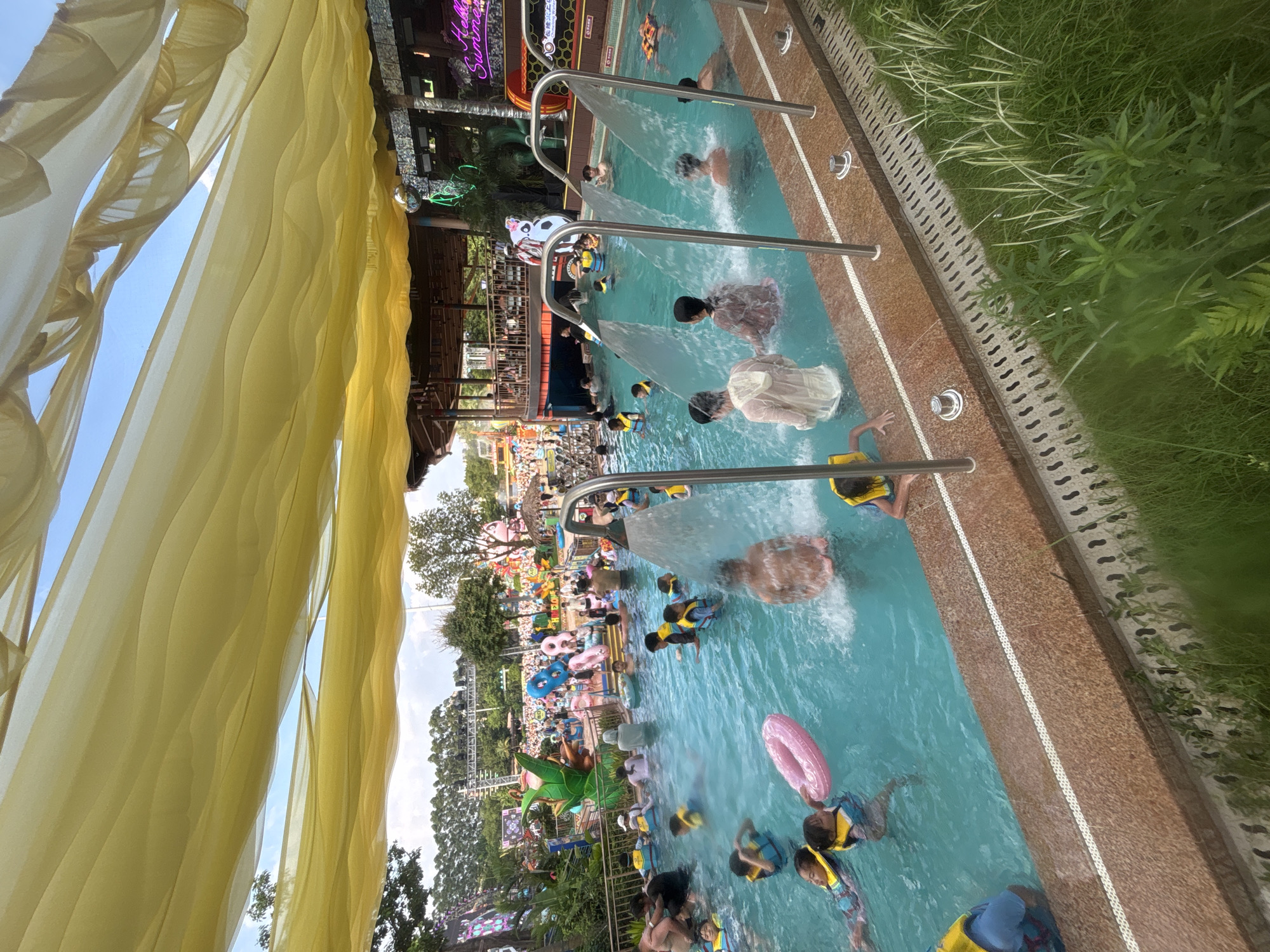 In Panyu District, China, a crowded swimming pool at a water park is depicted under a yellow, billowing canopy. Numerous individuals, including children wearing life vests, are submerged in the turquoise water. Several people are under streams of water cascading from shower-like fixtures. In the background, a multi-story structure with open-air seating and brightly colored decorations is visible. The scene suggests a lively recreational environment, likely during warm weather.