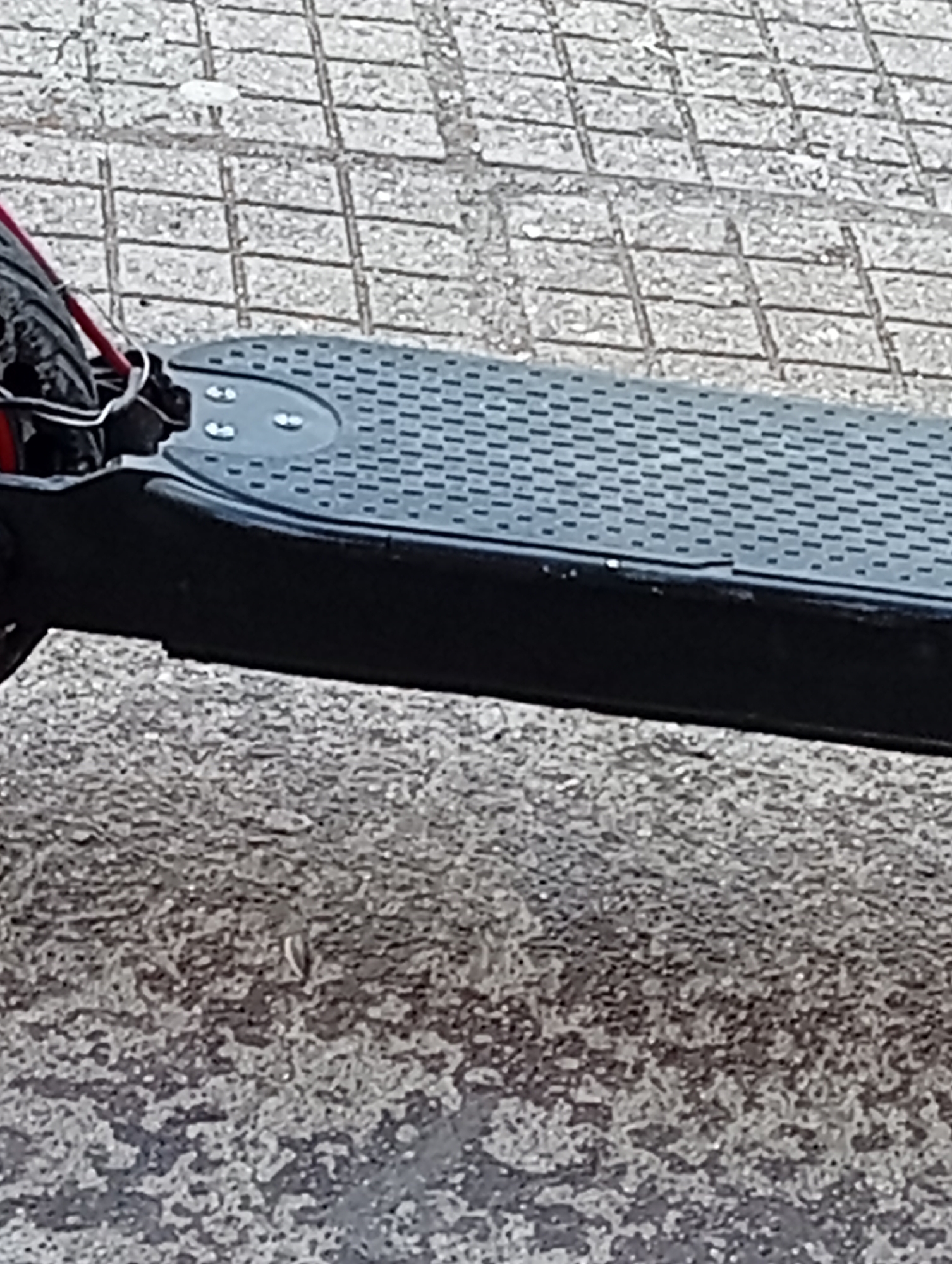 The image shows a close-up of an electric scooter, primarily focusing on the footboard. The footboard is black with a textured grey surface. The front of the scooter, including the tire and some wiring with red casing, are visible in the upper left. The scooter is positioned on a paved surface. The upper portion of the image features square paving stones, while the lower section displays a rougher, asphalt-like surface. The location is unspecified within Malko Chochoveni, Bulgaria.