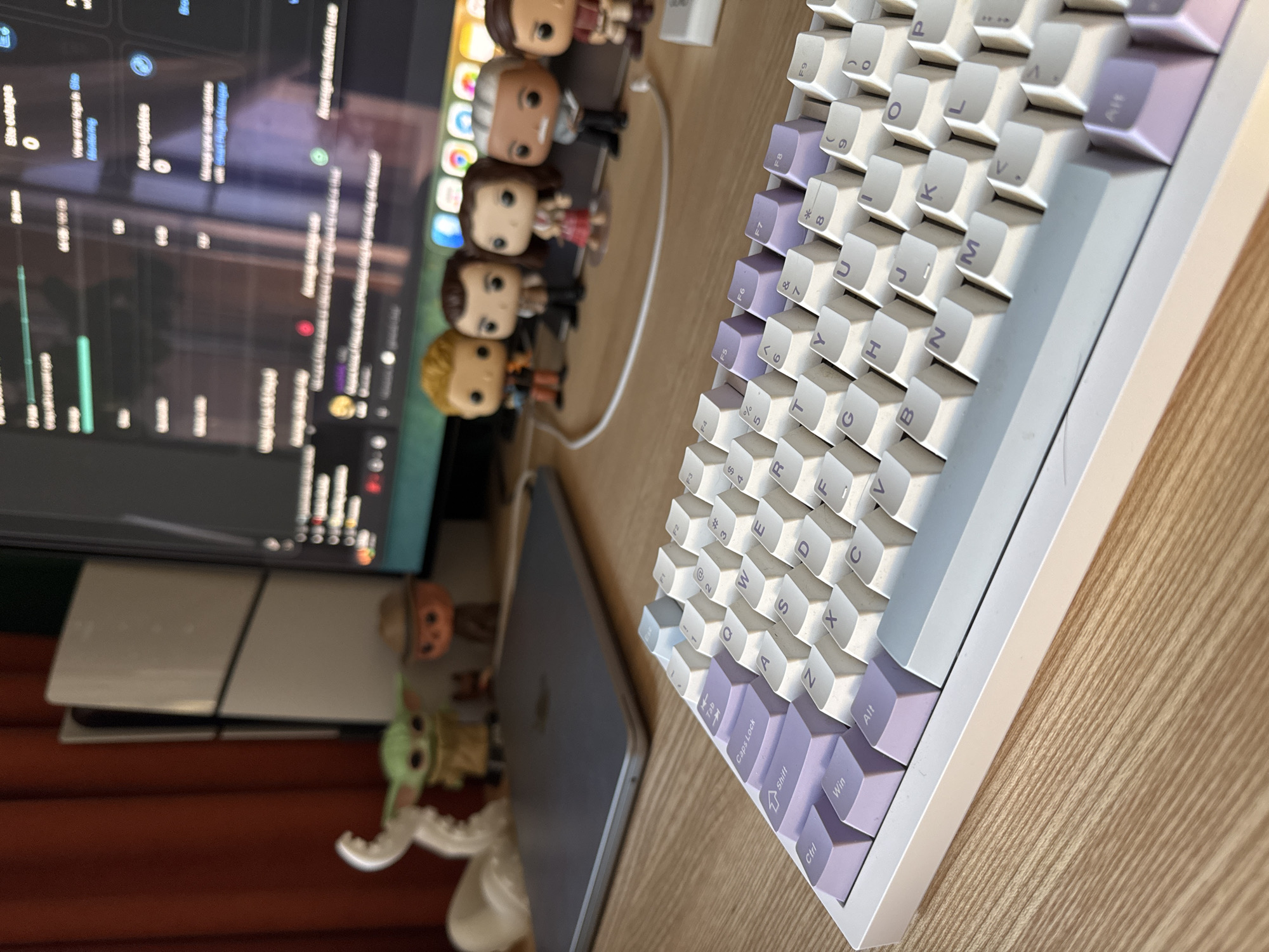 This is a close-up, eye-level shot of a white keyboard with purple and light blue keycaps, placed on a wooden desk. The keyboard is positioned in the foreground, taking up the majority of the lower half of the frame. Behind the keyboard, a laptop is visible, closed and silver. Further in the background, a computer monitor displays code or data, and several small figurines, including characters from Star Wars and other pop culture, are arranged on the desk. To the left of the monitor, a white vertical object, possibly a game console, stands against a red curtain. The lighting is natural, suggesting an indoor setting. The image does not provide any information that would allow for its location to be identified as Bila Tserkva, Ukraine.