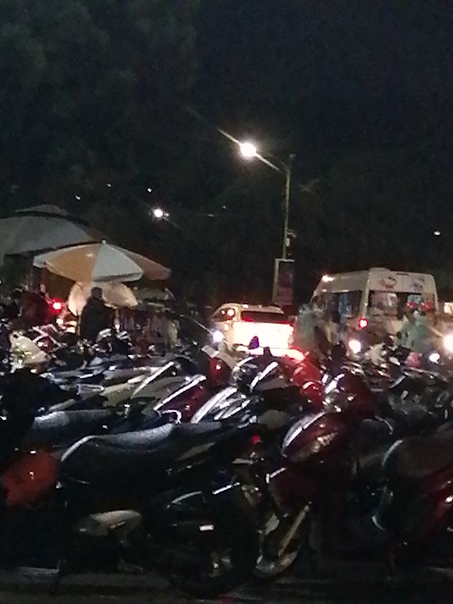 The image depicts a nighttime scene in Da Lat, Vietnam. A crowded motorcycle parking area is in the foreground, filled with numerous motorbikes. Overhead, there are market umbrellas covering vendor stalls where people are congregating. The background features a street with a parked white van and a light pole.