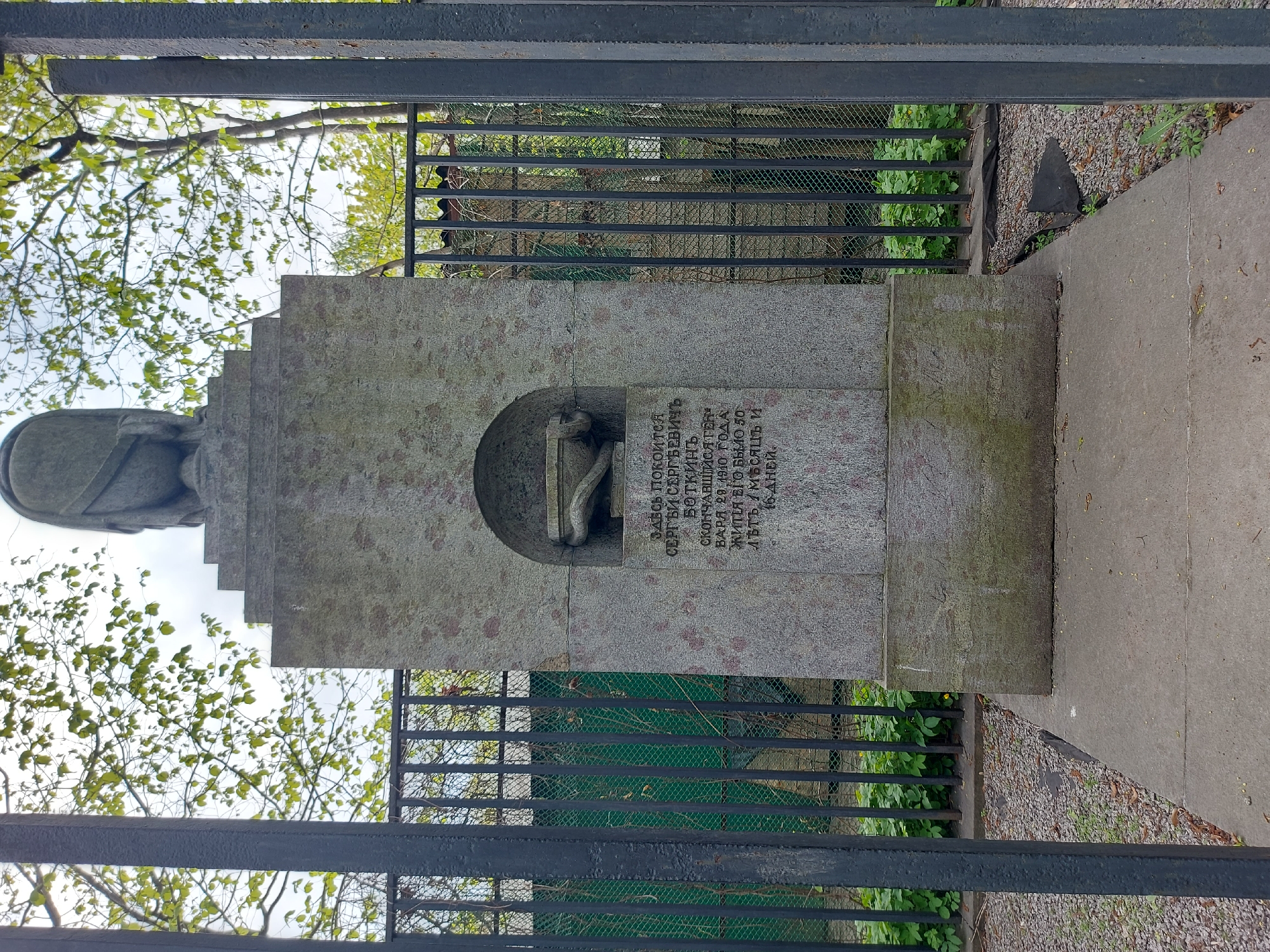 The image depicts the tombstone of Sergei Sergeevich Botkin located in Saint Petersburg, Russia. The stone is a large, rectangular structure with a semi-circular niche containing a small sculpture. An inscription in Russian is visible on the stone. The memorial is enclosed by a black metal fence. The background includes foliage and other vegetation. The photo is taken from a slightly elevated perspective through the fence.