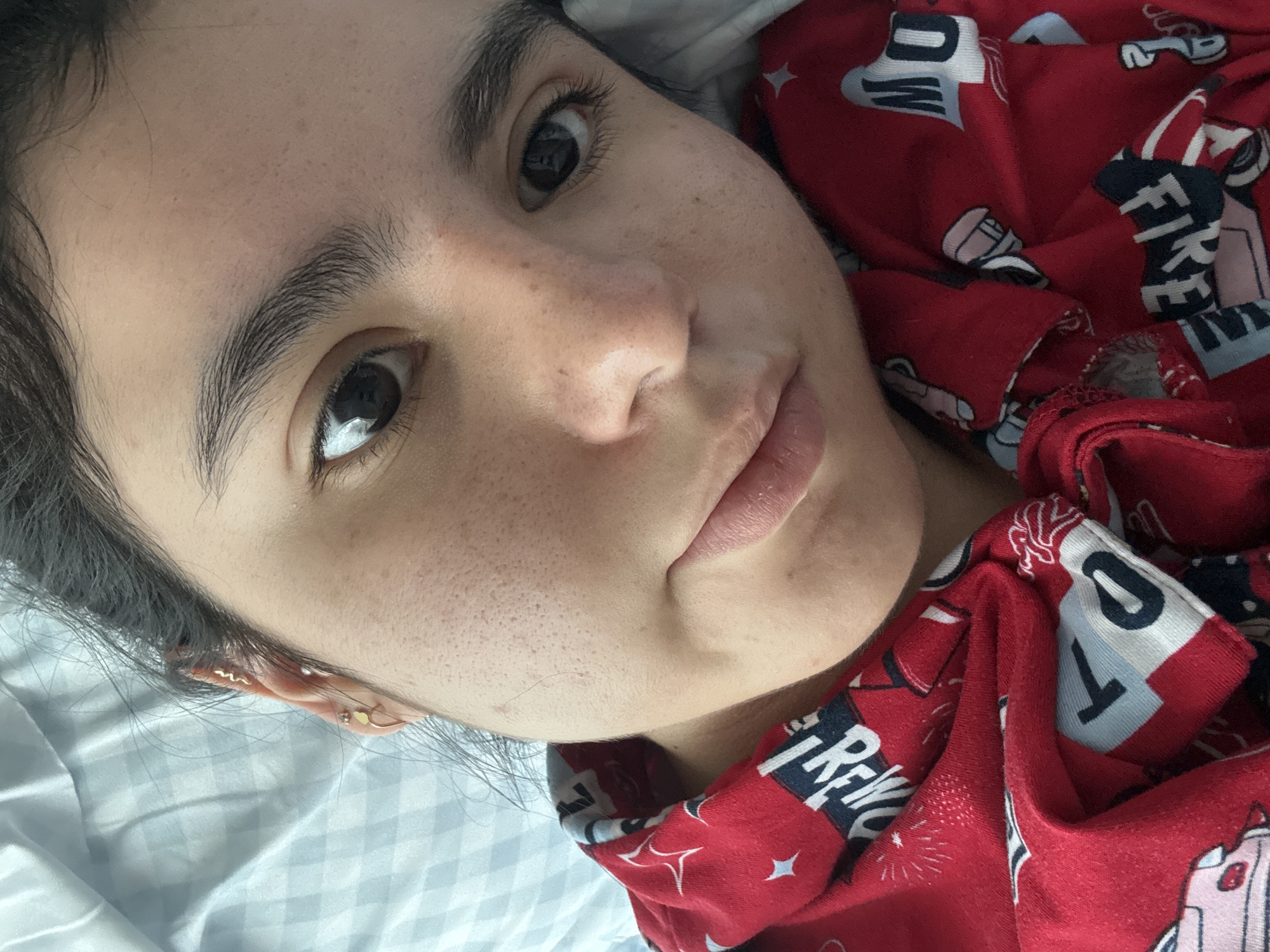 A close-up, high-angle portrait features a young adult with fair skin and dark hair, lying down and looking directly into the camera. The subject's head is tilted, resting on light-colored, possibly white or light blue, checkered bedsheets. Their dark hair is mostly pulled back, with a few strands visible around the face and ear.
Notable facial features include dark, almond-shaped eyes with visible eyelashes, and thick, well-defined dark eyebrows. The skin shows visible pores and a scattering of freckles across the nose and cheeks. The lips are full with a neutral, relaxed expression.
The person is wearing a red garment, likely pajamas, which has a distinct pattern. The pattern features various elements related to firefighting, including stylized fire trucks, fire extinguishers, and bursts resembling fireworks. Visible text on the fabric includes the word "FIRE" and the partial word "POW" (or "OW").
On the visible ear, multiple small, delicate gold-toned earrings are present: one shaped like a wavy line, a small heart stud, and another simple stud or hoop. The overall mood is relaxed and candid. The image is set in Austin, United States of America.