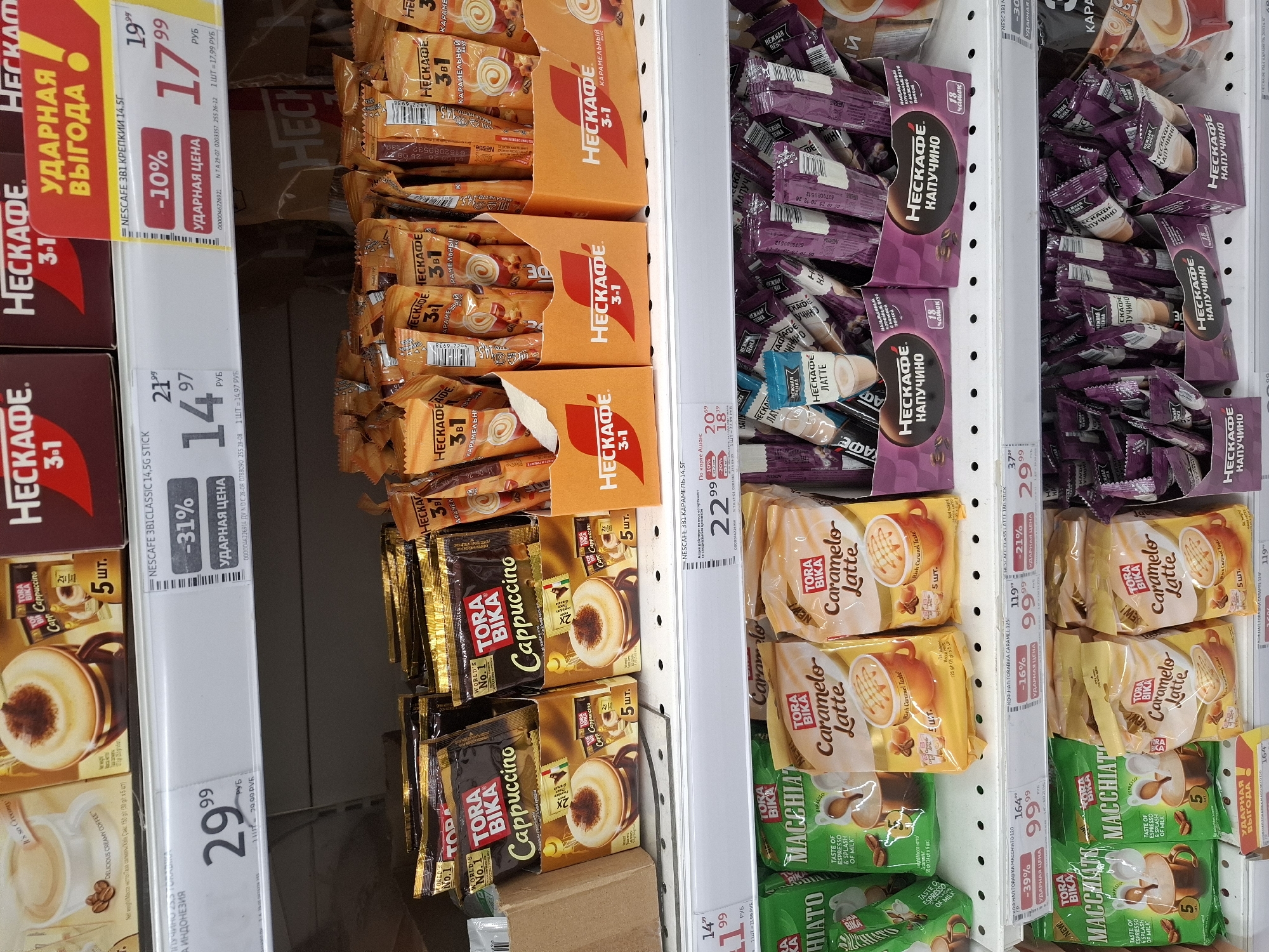 The image is a shelf display of instant coffee products in a retail environment, likely a supermarket or convenience store. The products are predominantly branded with "NESCAFÉ" and "TORA BIKA", featuring varieties such as Cappuccino, Caramel Latte, Macchiato, and "3-in-1" blends. The packaging is colorful, with prominent use of orange, brown, and purple. Many items are displayed in cardboard tiered stands.
Pricing and discount labels are visible on the shelves. For example, one label indicates a discount of "-31%" on "NESCAFÉ 3B1 CLASSIC 14,5G STICK", with a price of 14.97 RUB. Another shows "-10%" for "NESCAFÉ 3B1 КРЕПКИЙ 14,5Г" at 17.99 RUB. Further down, a "TORA BIKA MACCHIATO" is discounted by "-39%" to 99.99 RUB.
The lighting suggests an indoor retail setting, with bright, even illumination across the shelves. The overall impression is one of a well-stocked aisle featuring a variety of coffee options. There are no people visible in the image.
Without additional information or context, it is impossible to determine the specific location within the city of Tyumen, Russia. The image is a generic representation of a retail product display.