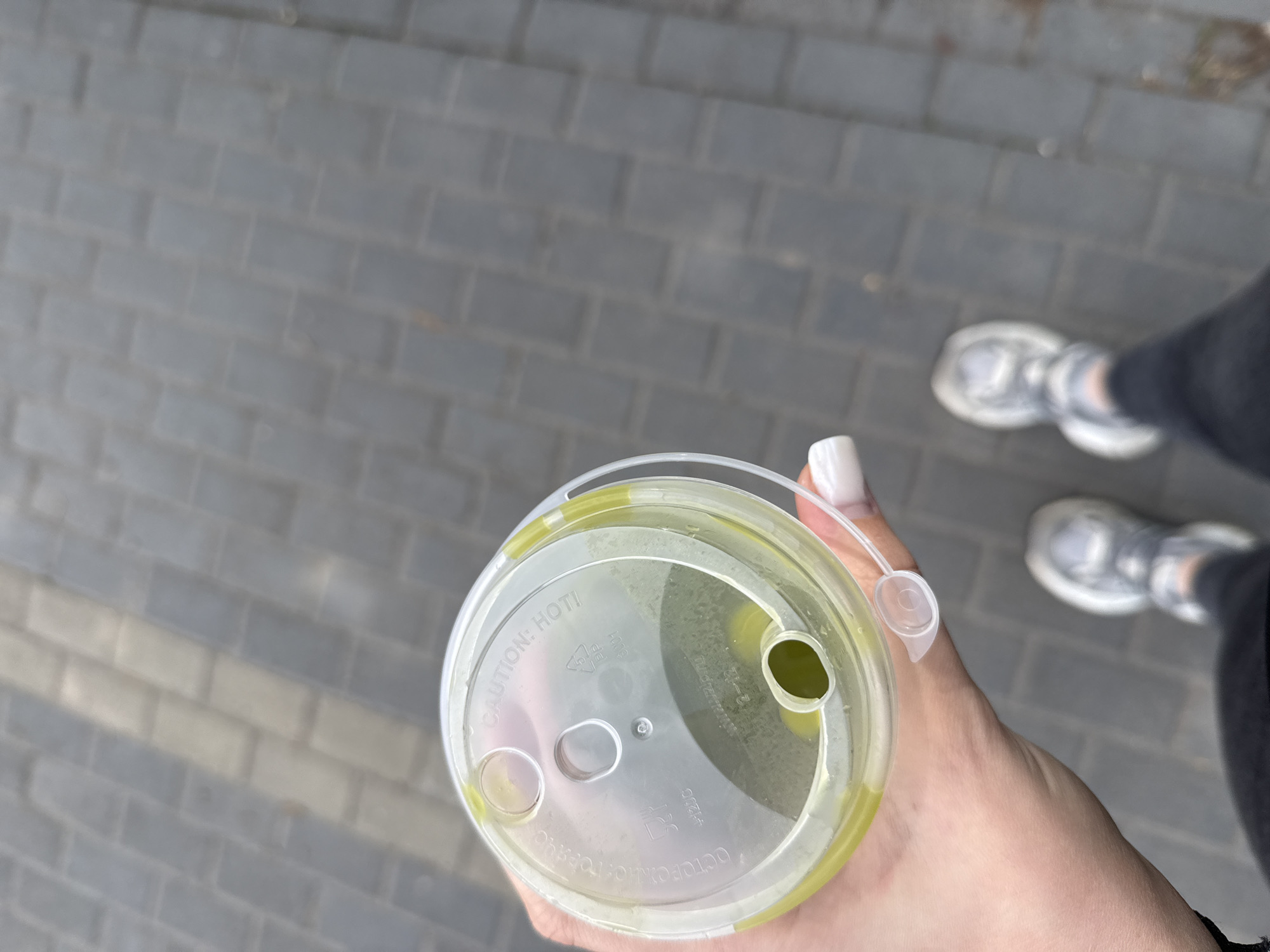 The image presents a first-person perspective of a person holding a clear plastic cup with a lid, containing a light green beverage, likely outdoors in Chaikovsky, Russia. The hand holding the cup has white-painted fingernails. In the slightly blurred background, a grey paver pathway is visible, along with the person's feet, clad in grey and white athletic shoes and dark pants, indicating a standing or walking posture. The scene is illuminated by natural light, suggesting it is daytime.
The transparent lid of the cup features several visible markings. Prominently displayed in English is the warning "CAUTION! HOT!". Adjacent to it, a recycling symbol is visible, alongside the Russian translation "OCTOPOЖHO: ГОРЯЧЕЕ" (Caution: Hot). Additional faint text and numerical symbols are also present on the lid. The lid has a large oval opening, presumably for a straw, and a smaller circular opening. The greenish liquid inside the cup appears to be a uniform color, possibly indicating a matcha drink or similar beverage.