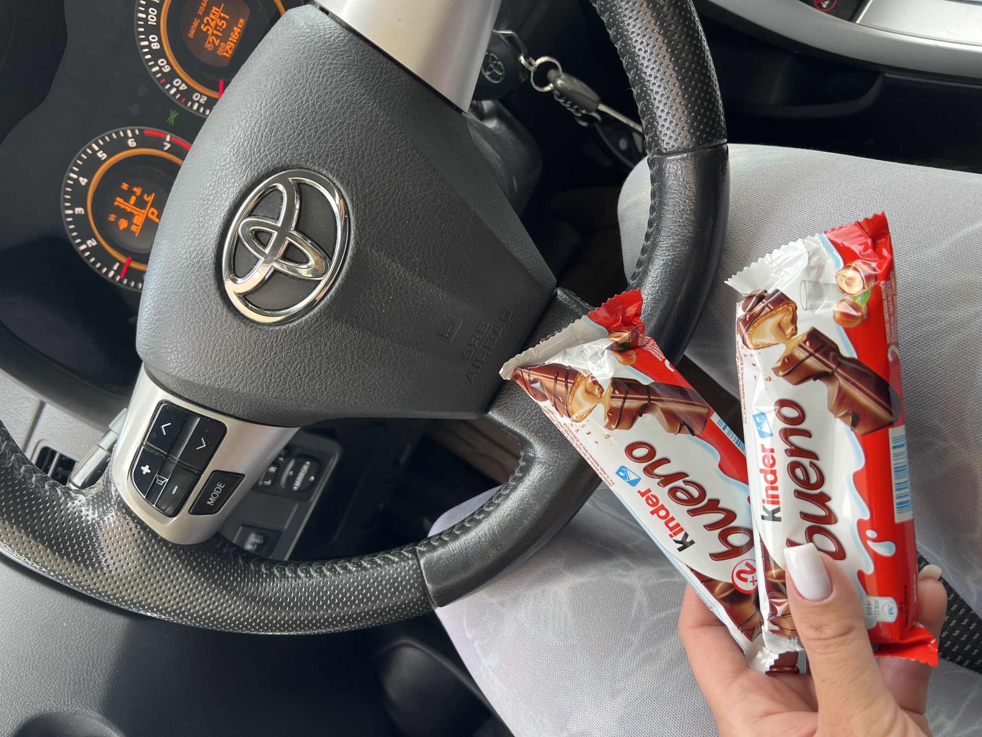 The image shows the interior of a car, specifically the steering wheel area of what appears to be a Toyota, judging from the car's logo. The dashboard is visible, displaying "129164 km" and "52 km". Two Kinder Bueno chocolate bars are held in a hand, placed in front of the steering wheel. The hand has white manicured nails. The setting seems to be within a vehicle, potentially parked or stationary, considering the focus on the interior and the presence of the chocolate bars. There is no specific information in the image that allows to pinpoint Budva, Montenegro as the location.