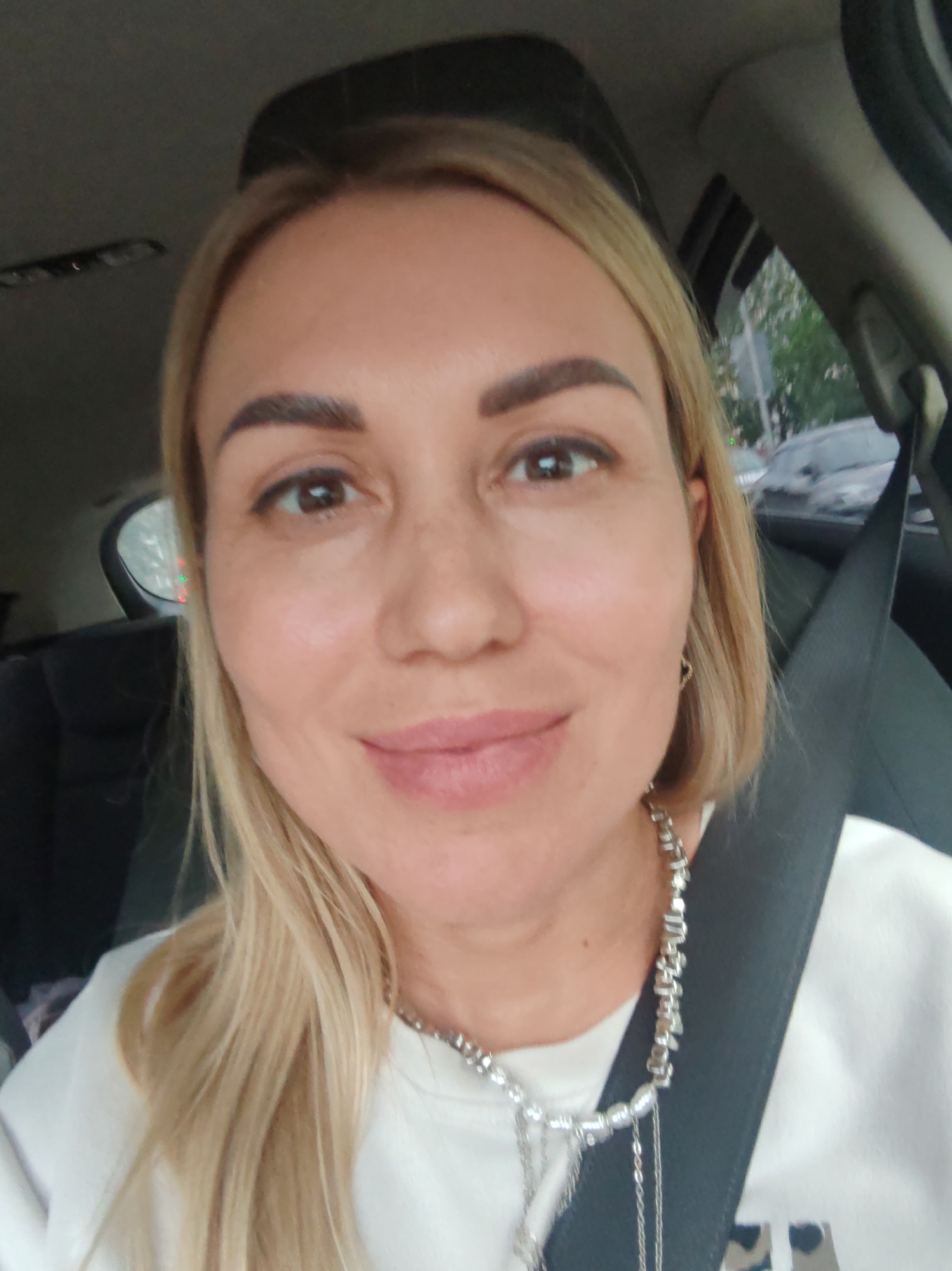 The image is a medium-shot, selfie-style photograph of a woman with blonde hair and brown eyes. She is in the driver's seat of a car, with her seatbelt fastened. The background shows the interior of the car and a blurred view of trees and cars outside the window. The woman is wearing a white sweatshirt and a silver necklace. She has a subtle smile and appears to be in a relaxed mood. The lighting suggests it is daytime, but there are no clear indicators of the weather or time of day beyond that. The location context is Komsomolsk-on-Amur, Russia. There is no visible text in the image.