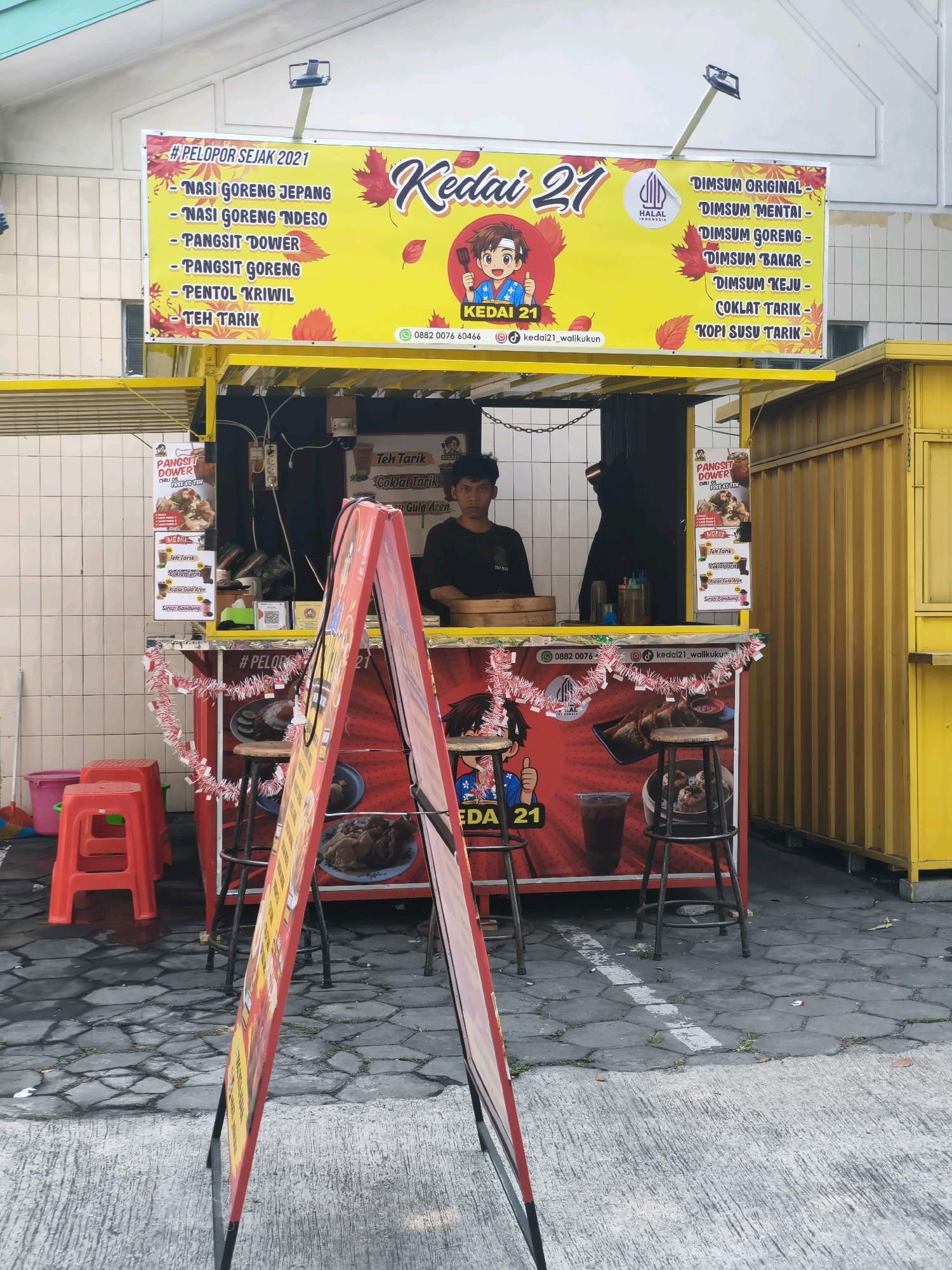A street food stall named "Kedai 21" is set up on a paved area with hexagonal stone tiles in Surakarta, Indonesia. The stall is adorned with a yellow banner featuring the establishment's name, a cartoon mascot, and a list of food and drink items, including "Nasi Goreng Jepang," "Dimsum Original," and "Teh Tarik." A young man with dark, curly hair is visible behind the counter, looking towards the camera. Red plastic stools are placed in front of the stall, and a red A-frame sign stands on the pavement. The stall is decorated with red and white festive garlands. A yellow corrugated metal structure is located to the right of the stall. The background shows a tiled wall and an outdoor environment.