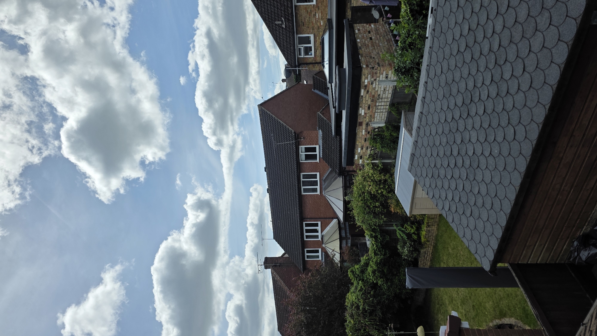 A view from above shows a suburban residential area under a partially cloudy sky. In the foreground, a dark, shingled roof of a wooden structure, possibly a shed or garage, dominates the bottom of the frame. Beyond this, a neatly mown green lawn is visible, with a tall, dark grey cylindrical object standing upright on the grass, which could be a decorative post or outdoor equipment. Several garden sheds, one light tan and another a muted grey, are situated further back.
Behind the sheds, a row of brick houses with dark tiled roofs is visible. The houses feature white-framed windows, some with internal blinds. Conservatories with angled, transparent roofs are attached to some of the houses. Lush green trees and dense foliage line the properties, creating a sense of a well-established garden environment. The sky above is a bright blue, interspersed with large, fluffy white cumulus clouds. The overall scene depicts a typical residential backyard in daylight, with a clear and somewhat overcast sky. There are no people or animals present in the image.