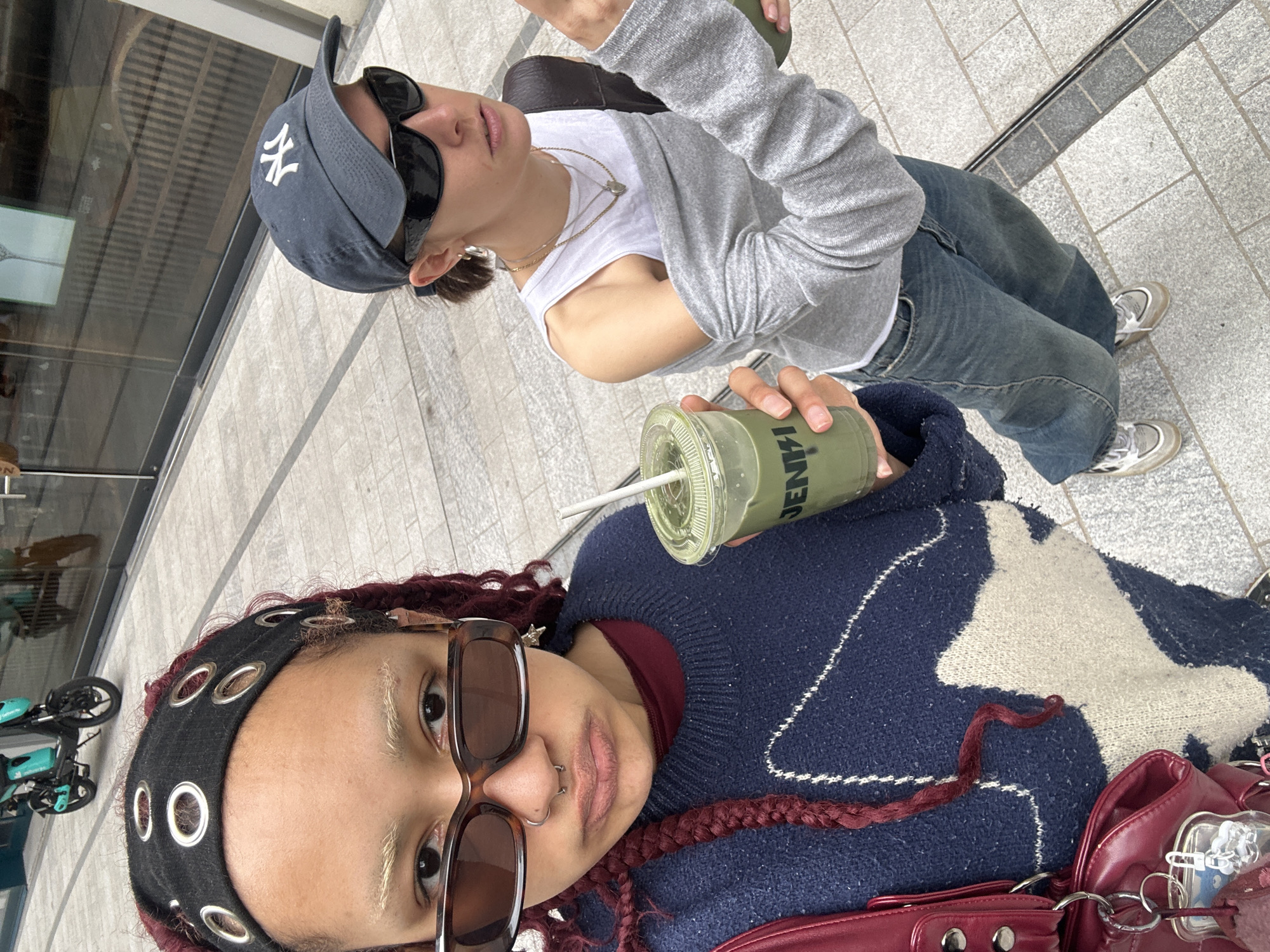 The image captures two individuals in an outdoor urban setting. The foreground person, with red braided hair and dark sunglasses, wears a black headband with circular metallic embellishments, a navy blue sweater with a light-colored abstract design, and carries a red shoulder bag. They hold a green-colored drink with a straw.
Behind them, a second person wears a New York Yankees baseball cap, dark sunglasses, a long-sleeved grey shirt over a white tank top, and blue jeans. They hold the same drink.
The background reveals a tiled pedestrian area, reflective glass buildings, and a parked teal-colored bicycle. Considering the architectural style and general ambiance, the location is likely in the city of London, United Kingdom.