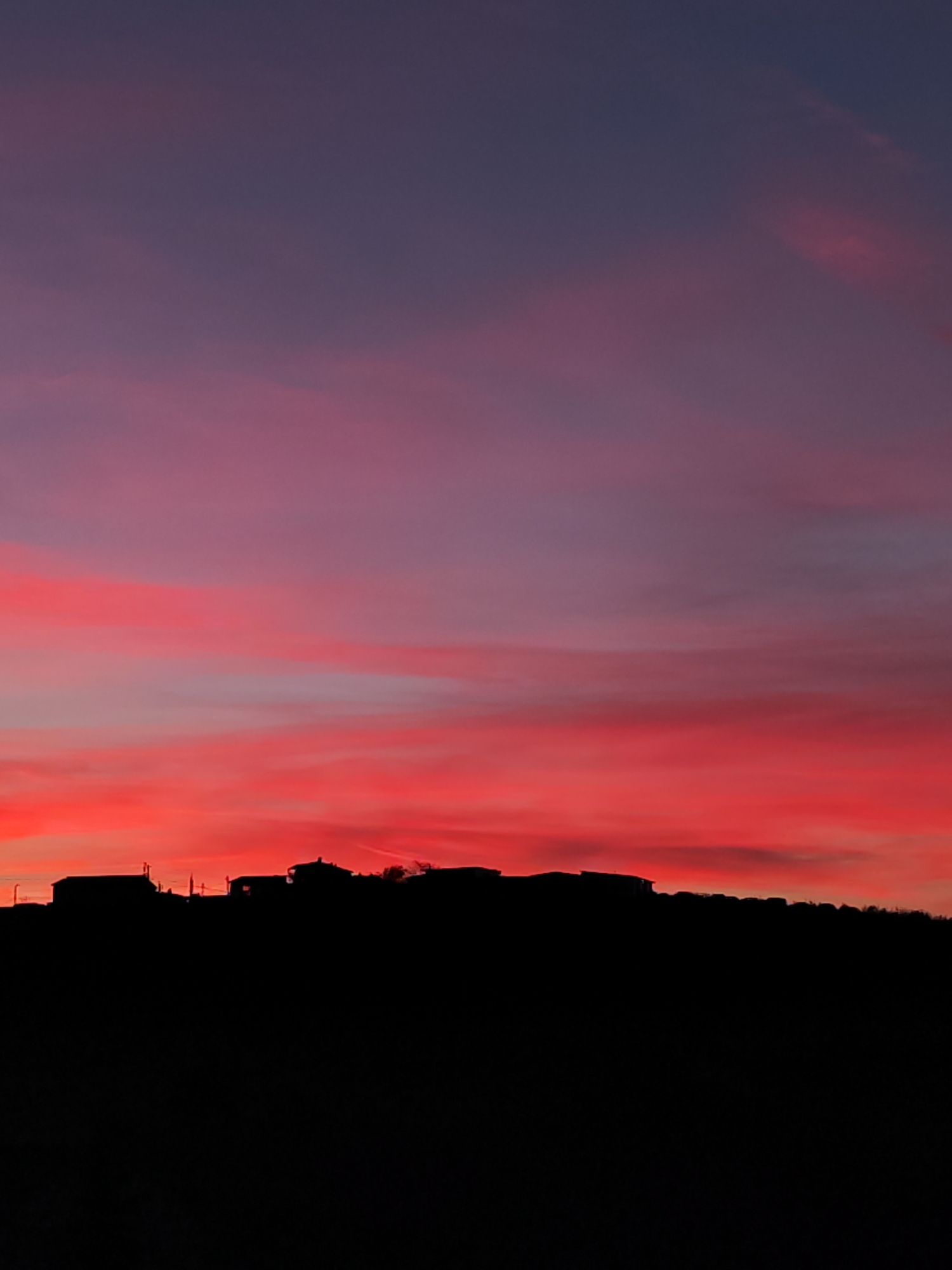 The image captures a striking sunset or sunrise over Burgas, Bulgaria, featuring a dramatic and colorful sky. The sky transitions from an intense, fiery red and orange at the horizon, where some elongated cloud streaks are visible, upwards into shades of deep purple and dark blue. The lower portion of the image presents a stark silhouette of the landscape, completely dark against the vibrant sky. This silhouette clearly outlines several buildings, likely houses, atop a hill or elevated terrain, along with the undulating line of the ground below them. No specific details of the buildings or landscape are discernible due to the silhouetted effect. The overall scene conveys a peaceful and awe-inspiring moment in nature, emphasizing the contrast between the brilliant sky and the shadowed earth. There are no visible people, specific activities, or discernible text in the image.