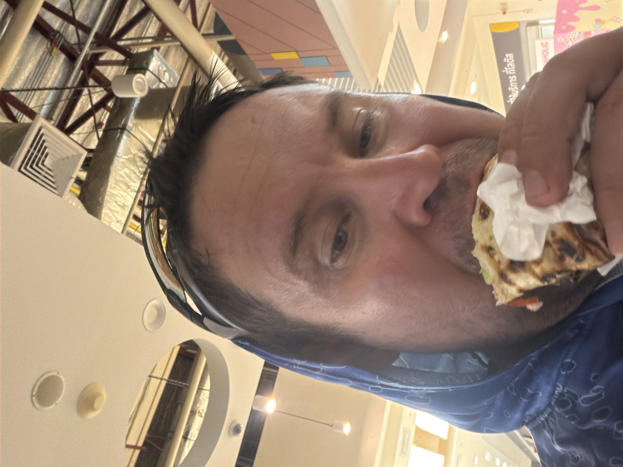 In the city of Ban Bang Rak, Thailand, a close-up, waist-up shot captures a man wearing a blue hooded sweatshirt with a patterned design and a black headband. He is in the process of eating a piece of food wrapped in white paper. The man is looking down at the food, with his mouth open as he takes a bite. The background shows a ceiling with exposed metal structures, lighting fixtures, and ventilation systems, typical of an indoor public space. There are also some signs visible in Thai script and a pink and yellow wall decoration. The overall lighting is bright and artificial.
