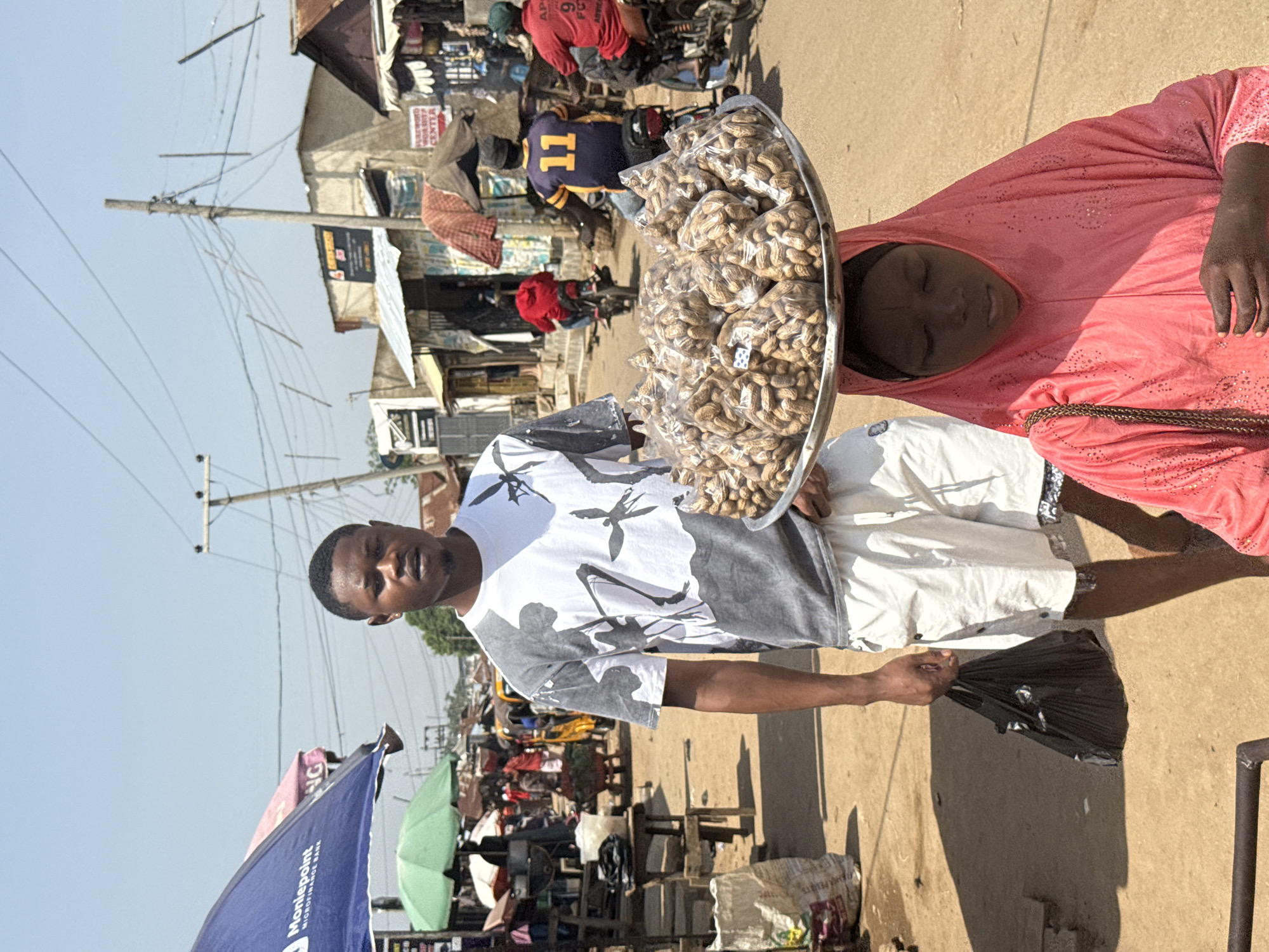 This media file depicts a lively outdoor market or street scene in Abuja, Nigeria, under bright daylight conditions.
In the foreground, two individuals are prominent. A man, wearing a white t-shirt with grey and black abstract designs, stands holding a black plastic bag in his right hand, looking towards the viewer. Beside him, a woman is seen carrying a large, metallic tray filled with numerous clear plastic bags of peanuts on her head. She is dressed in a vibrant pink, long-sleeved outfit that also covers her head, and her eyes appear closed or heavily lidded.
The background reveals a bustling environment with various elements. There are simple buildings, market stalls, and numerous utility poles with a dense network of electrical wires against a clear, bright blue sky. Several other people are visible, including individuals on motorcycles and others moving through the scene. One person in the mid-ground wears a blue and yellow jersey with the number "11" prominently displayed on the back. Another individual in a red shirt has "AFRICA" and "FOOTBALL" partially visible.
Notable details include a blue umbrella in the lower left corner bearing the text "Montepoint MICROFINANCE BANK". Further into the background, a sign for a "COMPUTER CENTER" can be seen. The strong shadows cast by people and objects indicate that it is daytime, likely mid-day, with sunny weather. The scene captures a typical street vendor and market interaction.