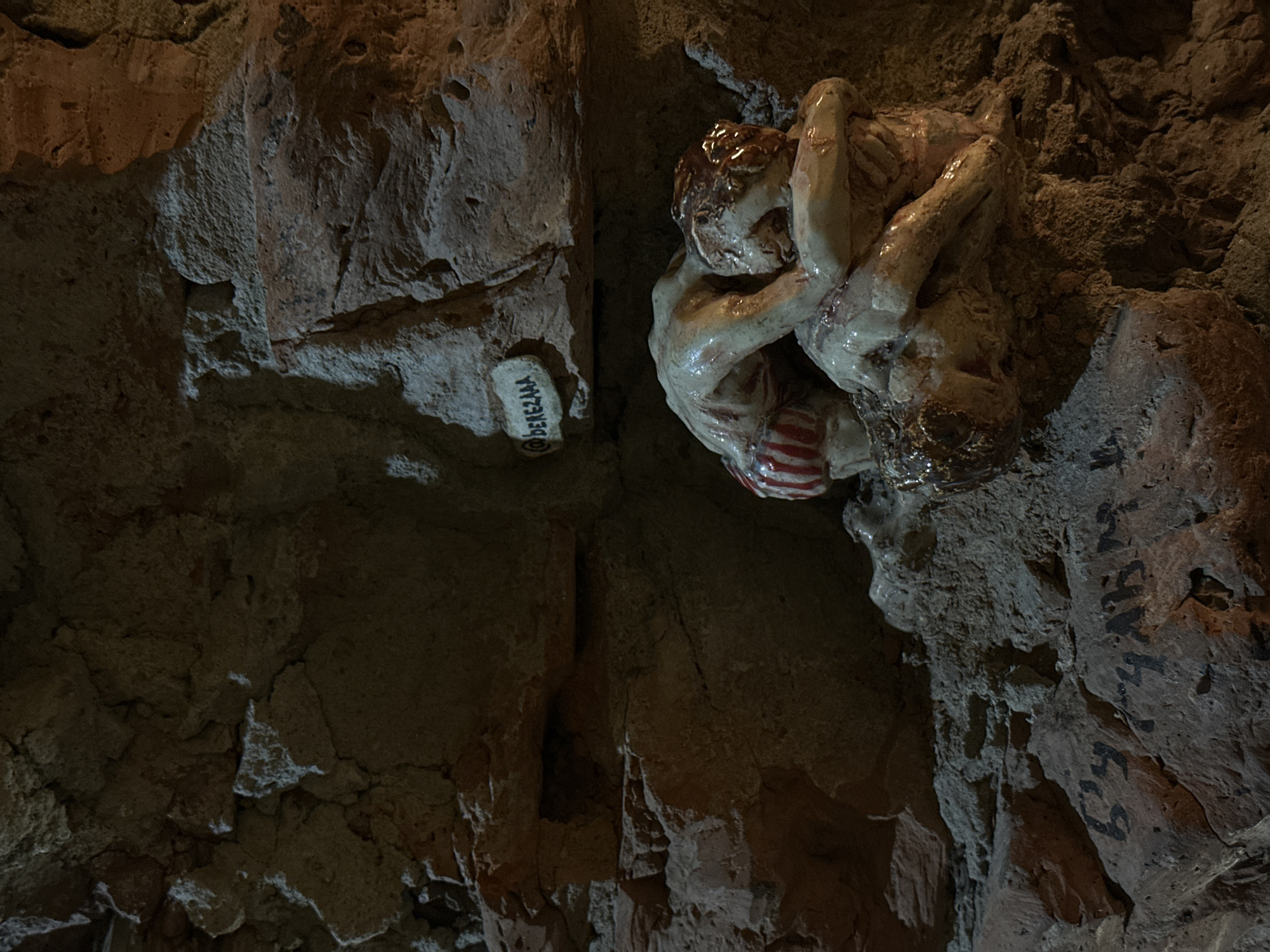 Here is a detailed description of the image:
The image depicts a close-up view of an artistic installation or sculpture within what appears to be a cave or grotto-like environment. The setting features rough, uneven rock surfaces with varying textures and a natural, rugged appearance.
The primary subject is a sculpture representing two nude figures intertwined in a struggle or embrace. The figures appear to be made of a glazed ceramic material, with details such as hair and musculature visible. One figure has red and white striped shorts. The sculpture is positioned on the right side of the image.
To the left of the figures is a small, white object with the name "DEREZAAA" printed in black lettering. The overall lighting is subdued, creating shadows and highlighting the textures of the rock and sculpture. The setting gives the impression of a historical or artistic site. No other people, animals, or specific actions are depicted in this static image.