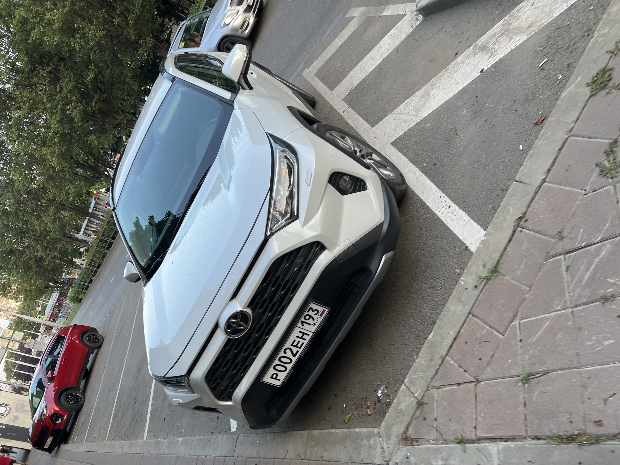 A white Toyota RAV4 is parked at an angle on a paved street in Krasnodar, Russia, in front of a sidewalk with red brick pavers. The car's license plate is visible and reads "P002EH 193 RUS". A red Kia Sportage is parked behind the Toyota. The street is lined with trees and buildings, and other vehicles are visible in the background. The scene appears to be in an urban setting during the daytime.