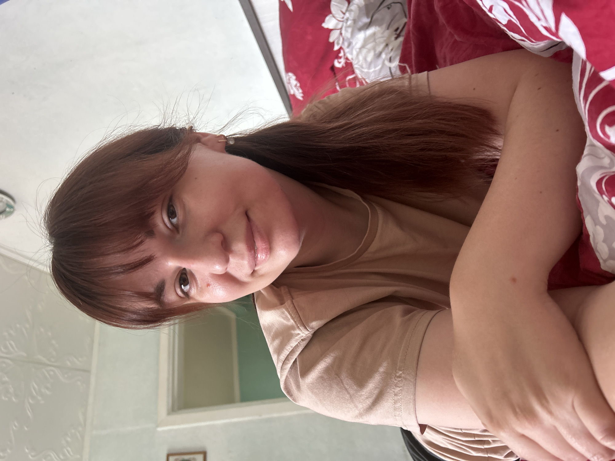 A woman with auburn hair and bangs lies on a bed covered with a red and white floral patterned duvet. She is wearing a light brown t-shirt and smiling slightly at the camera. The wall behind her is painted in a pale green color. A window and a framed picture are visible on the wall. The location Фоки, Russia cannot be determined from the image alone.