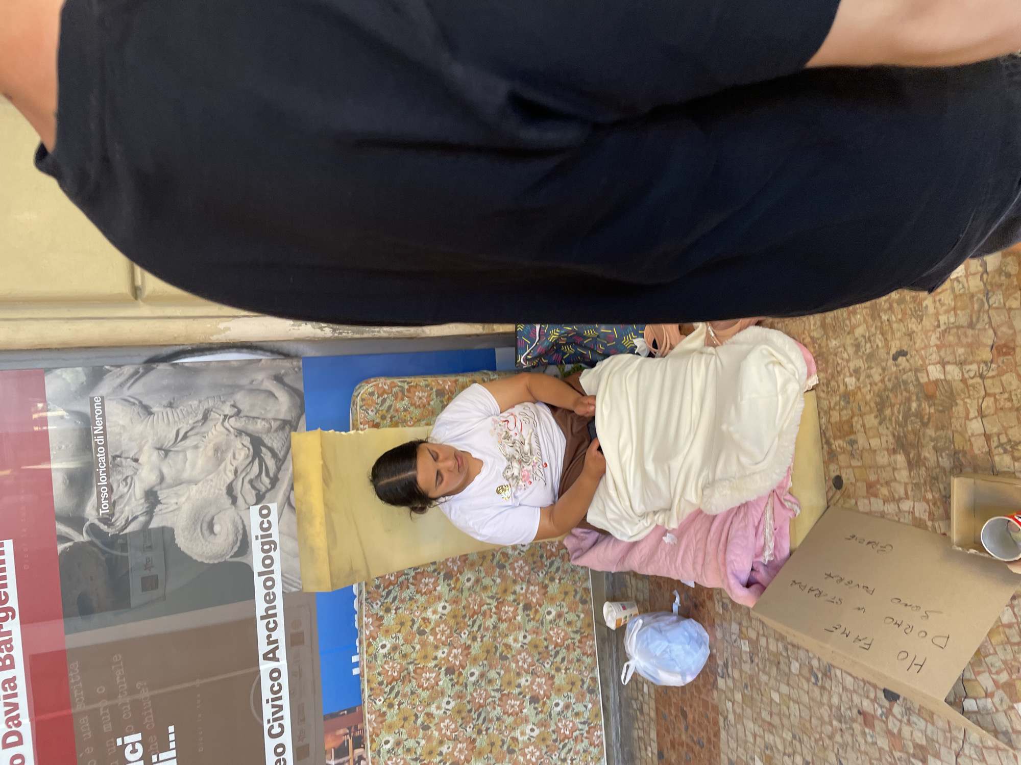 A woman lies on a makeshift bed on the ground next to posters in Bologna, Italy. The woman is resting on a mattress, covered with blankets. Next to her is a sign that reads "Ho fame, dormo in strada, sono povera" (I am hungry, I sleep on the street, I am poor) along with a donation cup. One poster refers to Torso loricato di Nerone (Torso clad in armor of Nero).