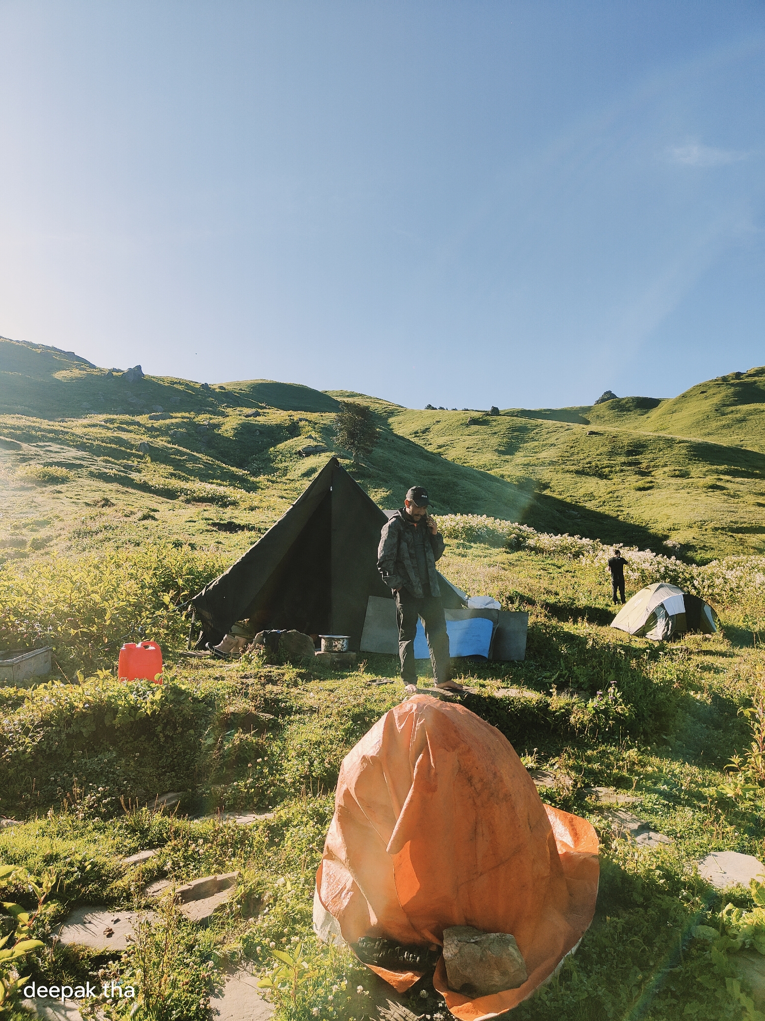 A group of tents are pitched on a grassy mountainside, with a bright blue sky overhead. In the foreground, a person wearing a dark jacket and a baseball cap is standing and talking on a cell phone, with a large orange tarp partially covering a pile of items beside them. To the left of this person, a dark green tent is pitched, with a red water jug and some camping gear nearby. Further back on the mountain, another person is standing near a small, light-colored tent. The landscape is covered in green grass and scattered white flowers, with rolling hills and rocky outcrops in the background. The image appears to have been taken in the morning or late afternoon, as the sun casts long shadows and illuminates the scene with a warm light. The overall impression is of a remote camping location, likely in the Himalayas near Manali, India, given the presence of the watermark "deepak.tha".